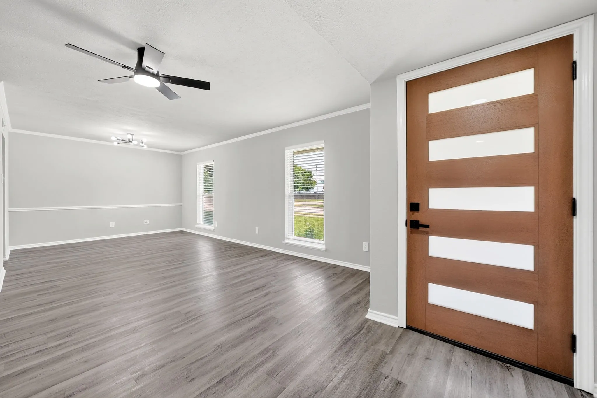 Modern front entry door with horizontal frosted glass inserts and matte black hardware, opening into an expansive room featuring wood-finish flooring, light gray walls with chair rail molding, and multiple windows with blinds