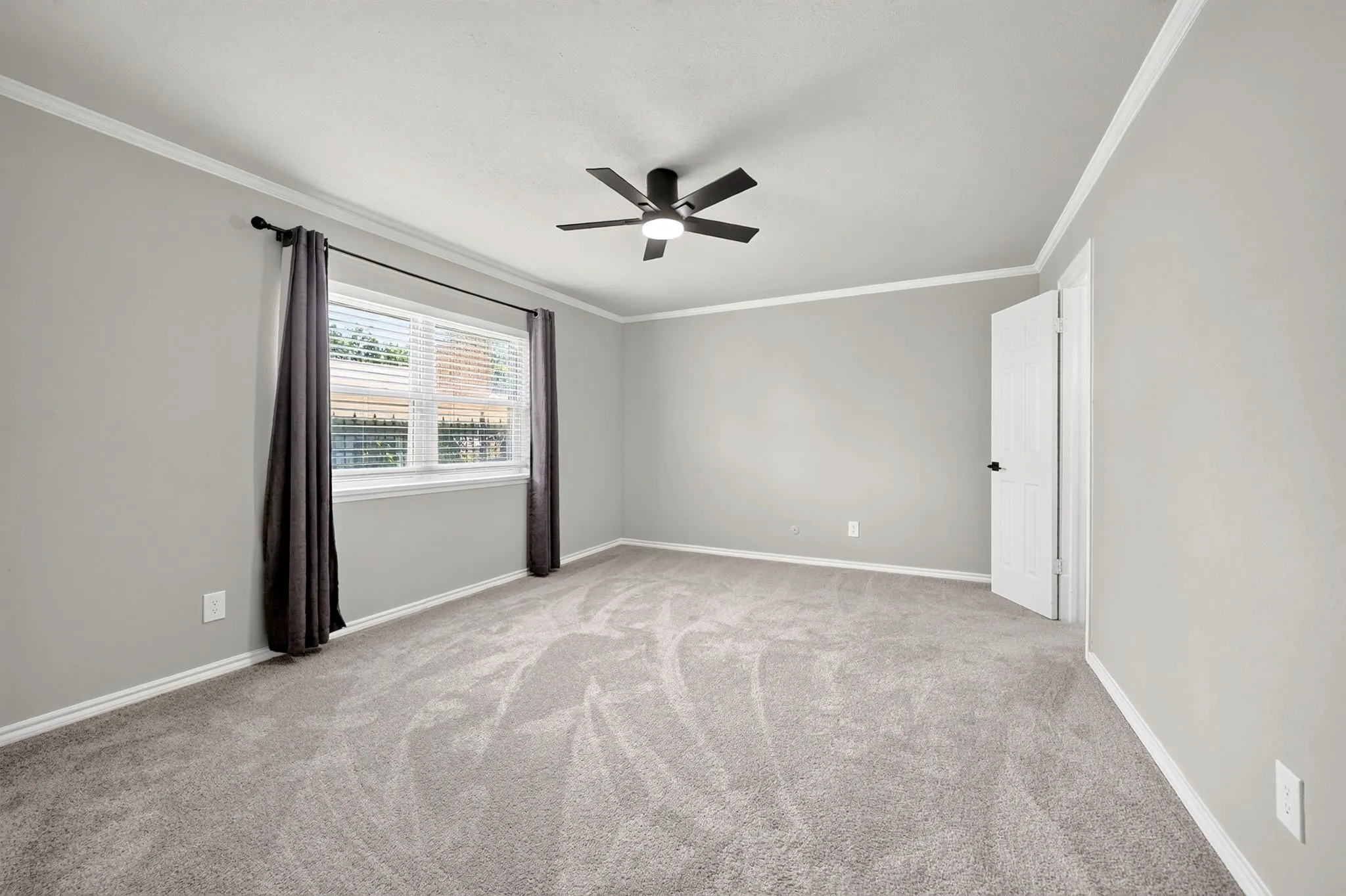 Spacious carpeted room featuring a ceiling fan with integrated lighting, a large window with blinds, white trim, and a paneled door with matte black hardware