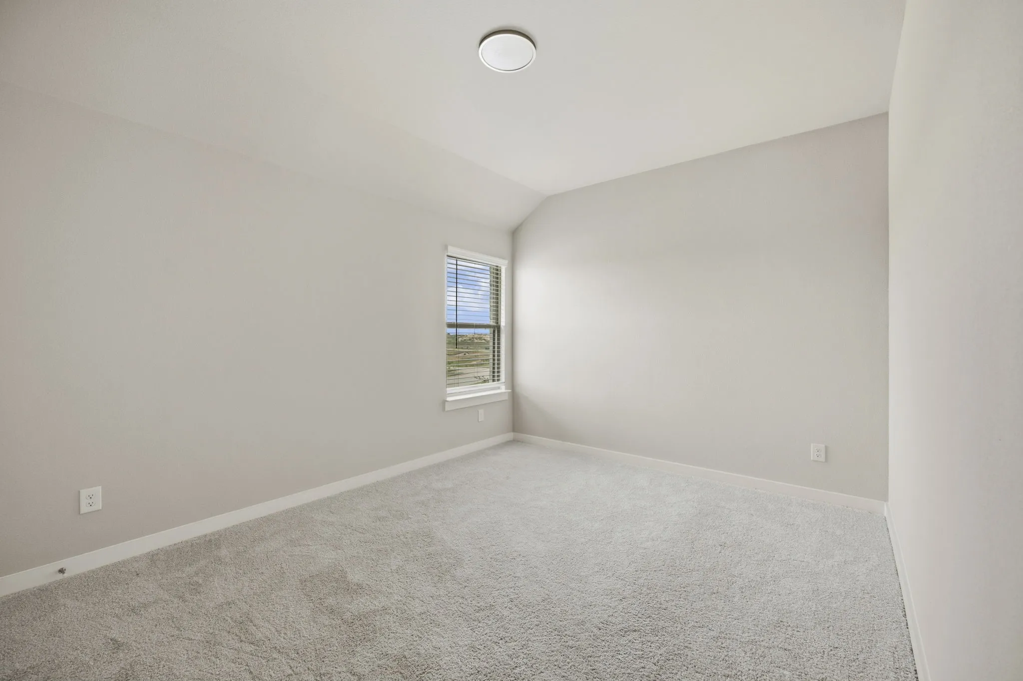 Spacious room featuring light-toned walls, plush gray carpeting, a single window with blinds, and a flush-mount ceiling light fixture