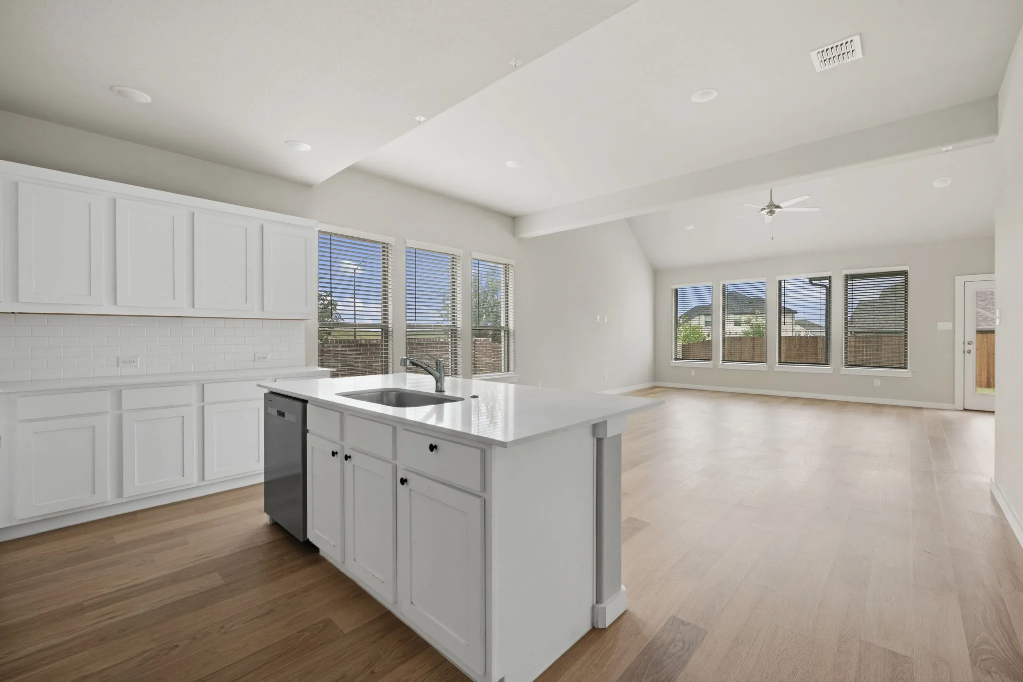 Spacious kitchen featuring white cabinetry, subway tile backsplash, and a central island with a sink, extending into an open living area with wood-finish flooring and vaulted ceilings