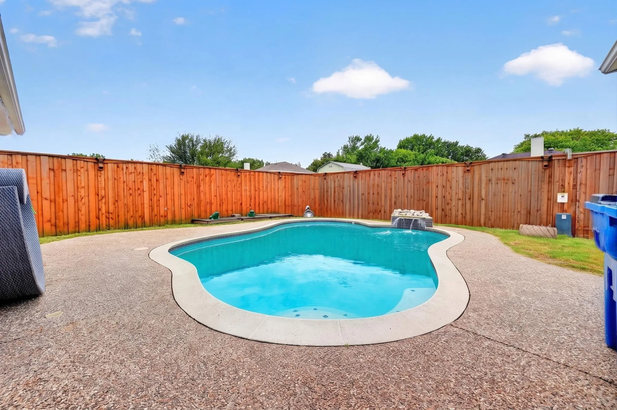 Curved inground pool with a waterfall feature, surrounded by an exposed aggregate patio and a wood privacy fence