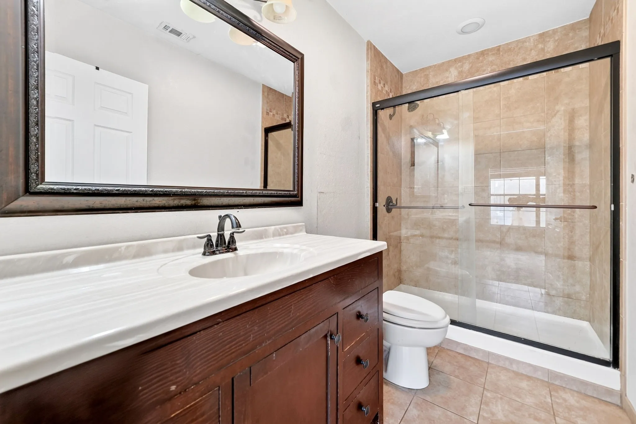 Bathroom featuring a dark wood vanity with a white countertop and integrated sink, a large framed mirror, and a walk-in shower with glass doors and tile surround