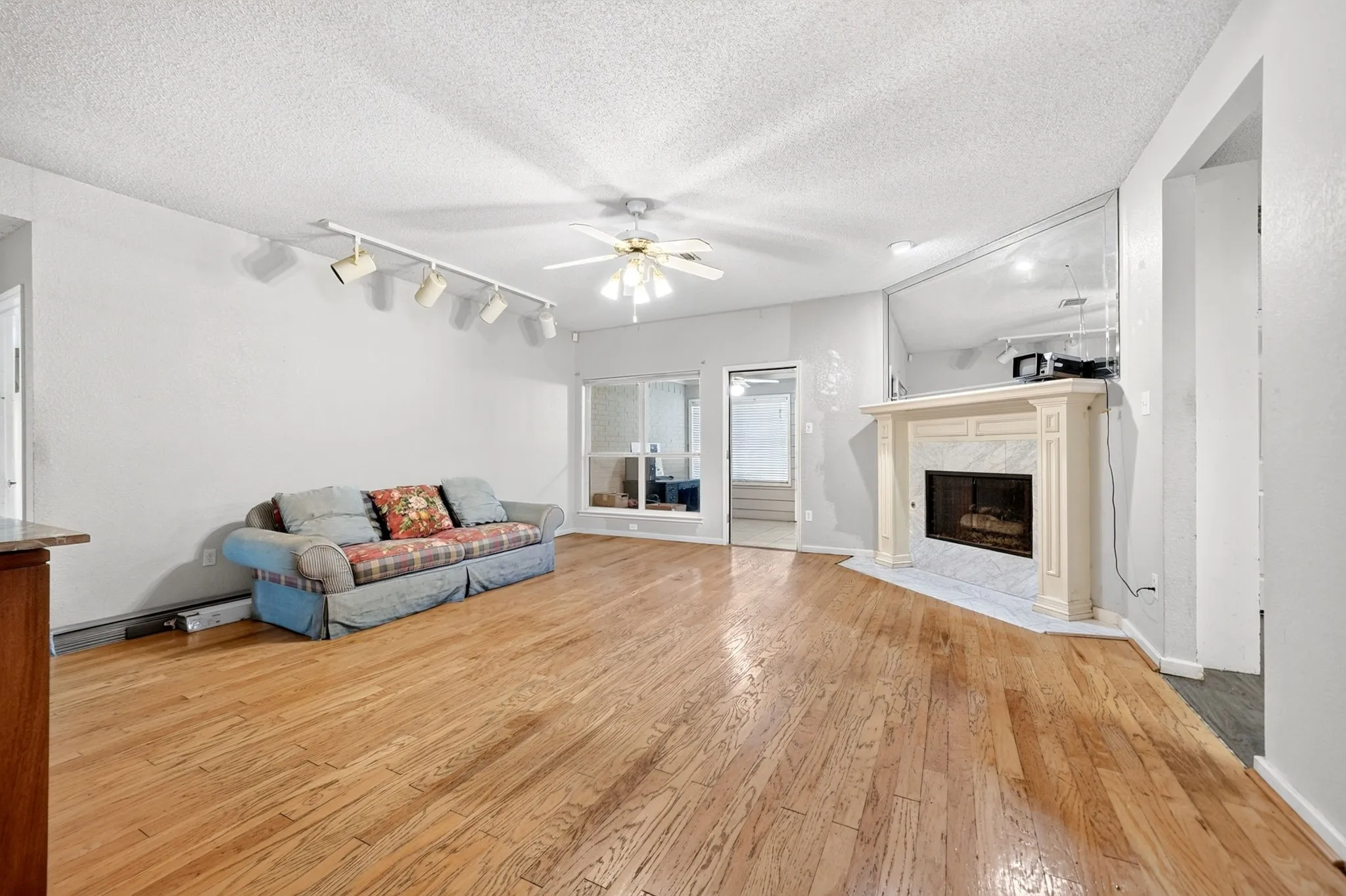 Spacious living area featuring wood flooring, a fireplace with a white mantel and tiled surround, track lighting, and a ceiling fan