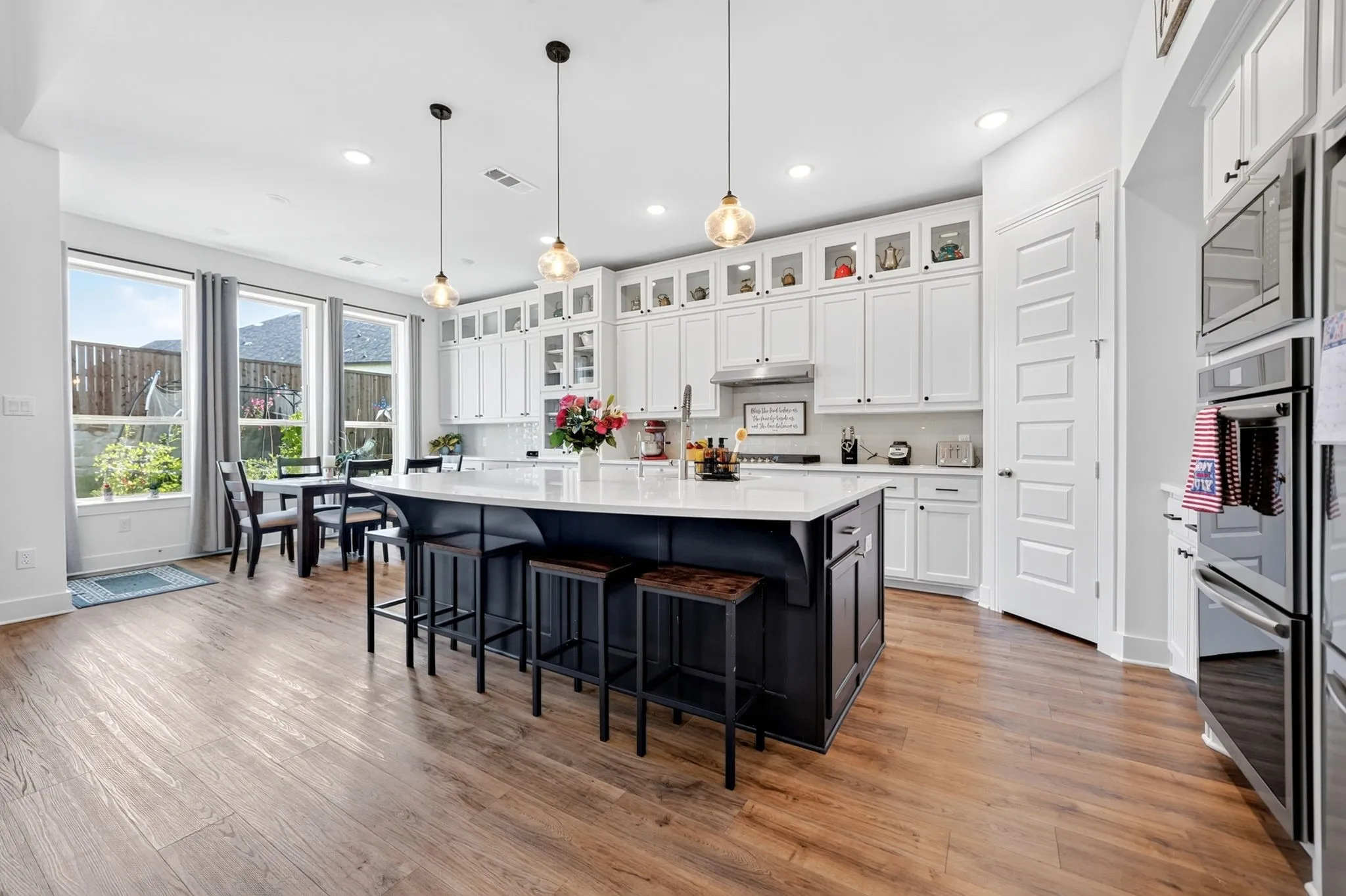 Spacious kitchen featuring a large island with seating, white shaker-style cabinetry with glass accents, stainless steel appliances, and wood-finish flooring