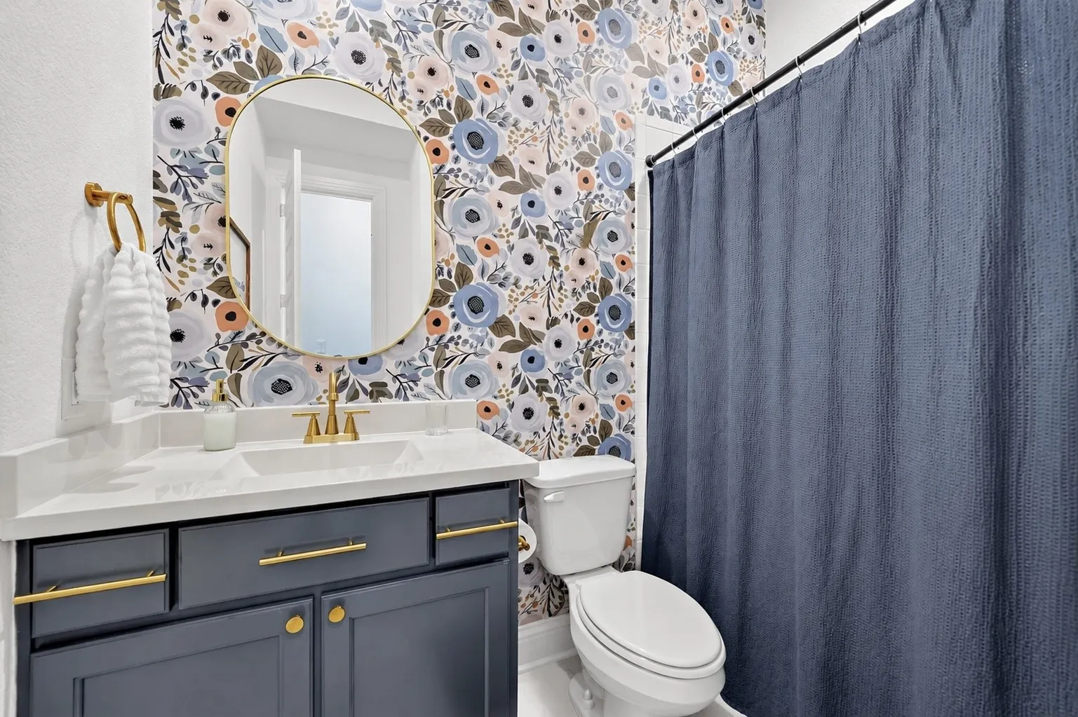 Bathroom featuring floral wallpaper, a white vanity with a rectangular integrated sink, a gold-framed oval mirror, a dark gray cabinet with gold hardware, and a white toilet
