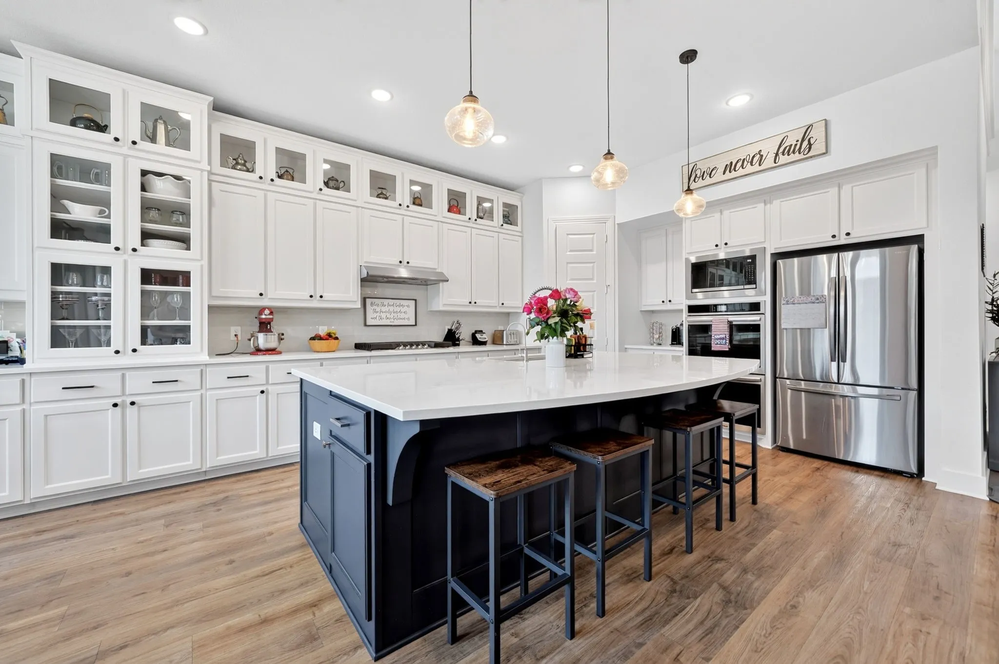 Modern kitchen featuring an expansive island with white countertop, white shaker cabinetry with glass-front uppers, stainless steel appliances, and wood-finish flooring