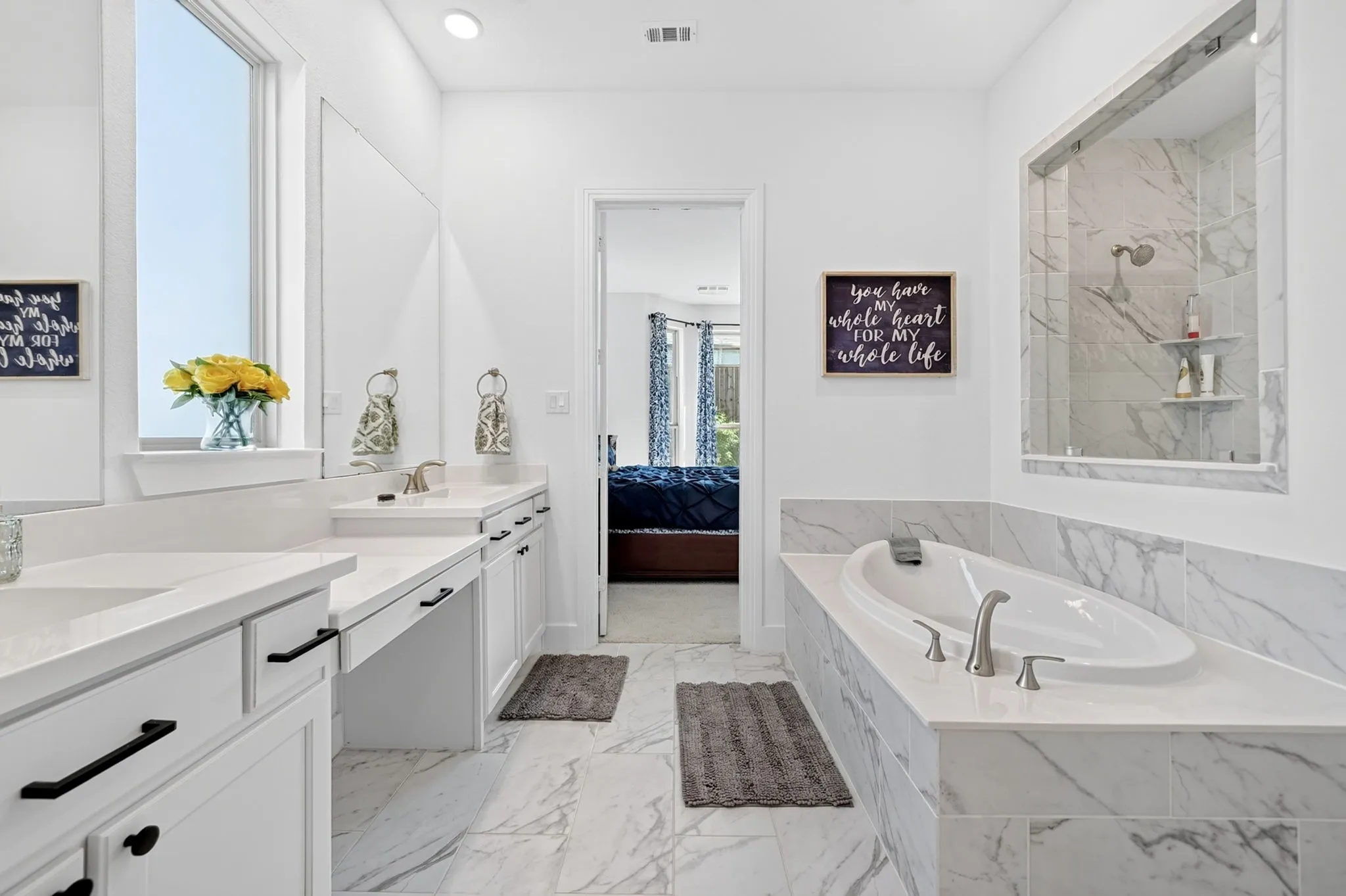 Spacious bathroom featuring white cabinetry with matte black hardware, dual vanity sinks, a built-in soaking tub with deck-mounted fixtures, and a glass-enclosed shower with a tiled surround