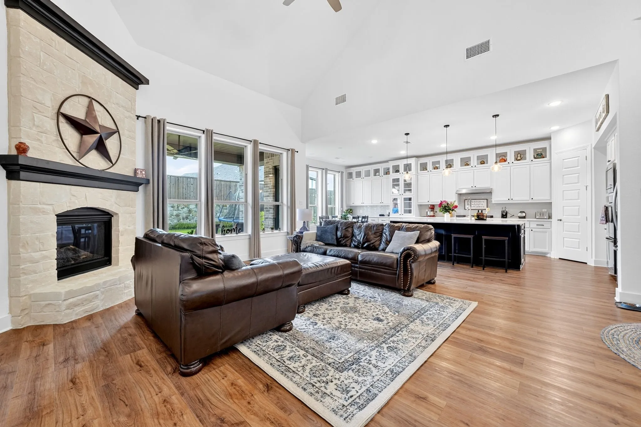 Spacious living area featuring a stone fireplace with a dark wood mantel, wood-finish flooring, and a bank of windows