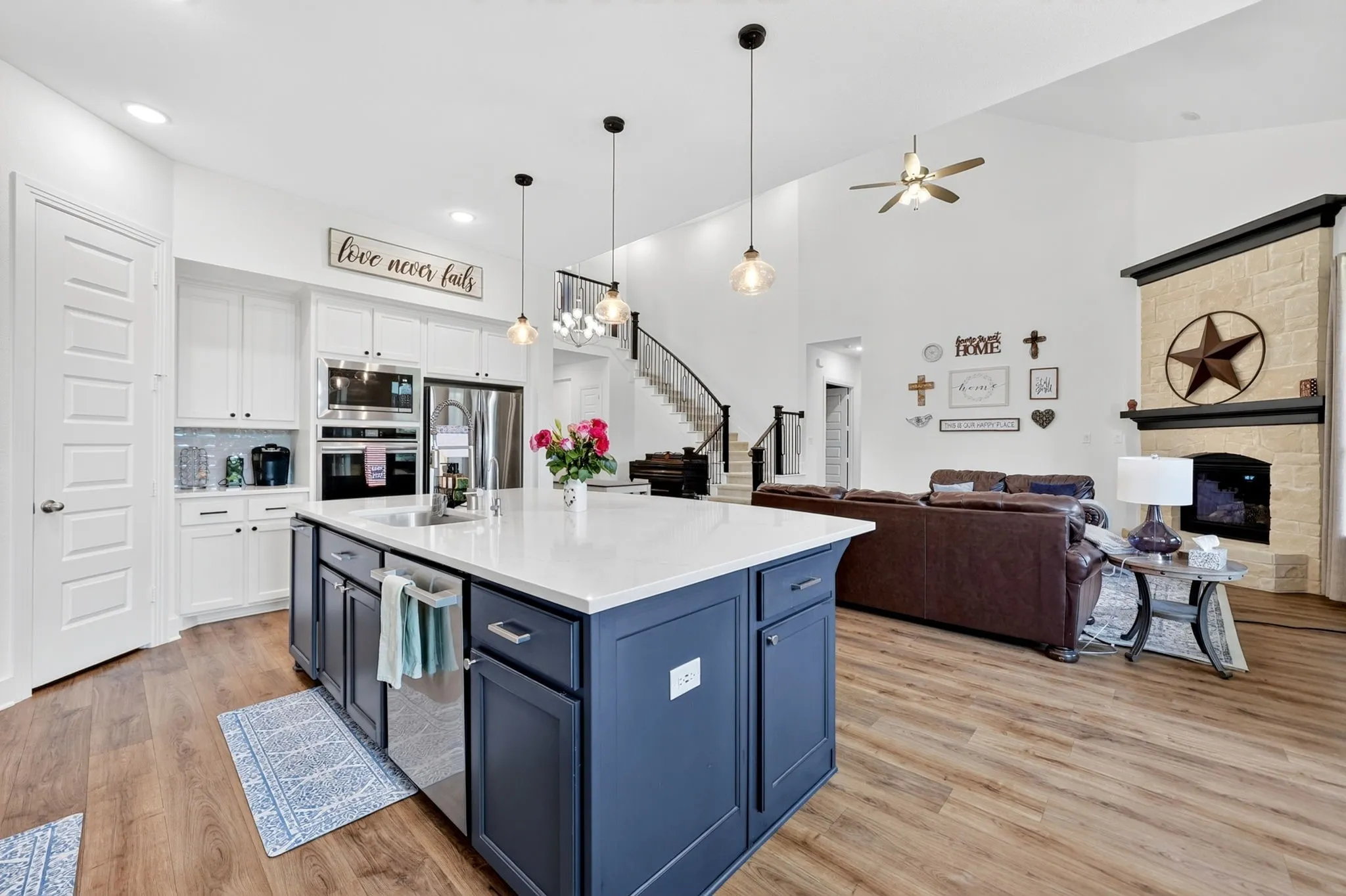 Open-concept living area featuring a kitchen island with pendant lighting, wood-finish flooring, a stone fireplace, built-in stainless steel appliances, and white cabinetry