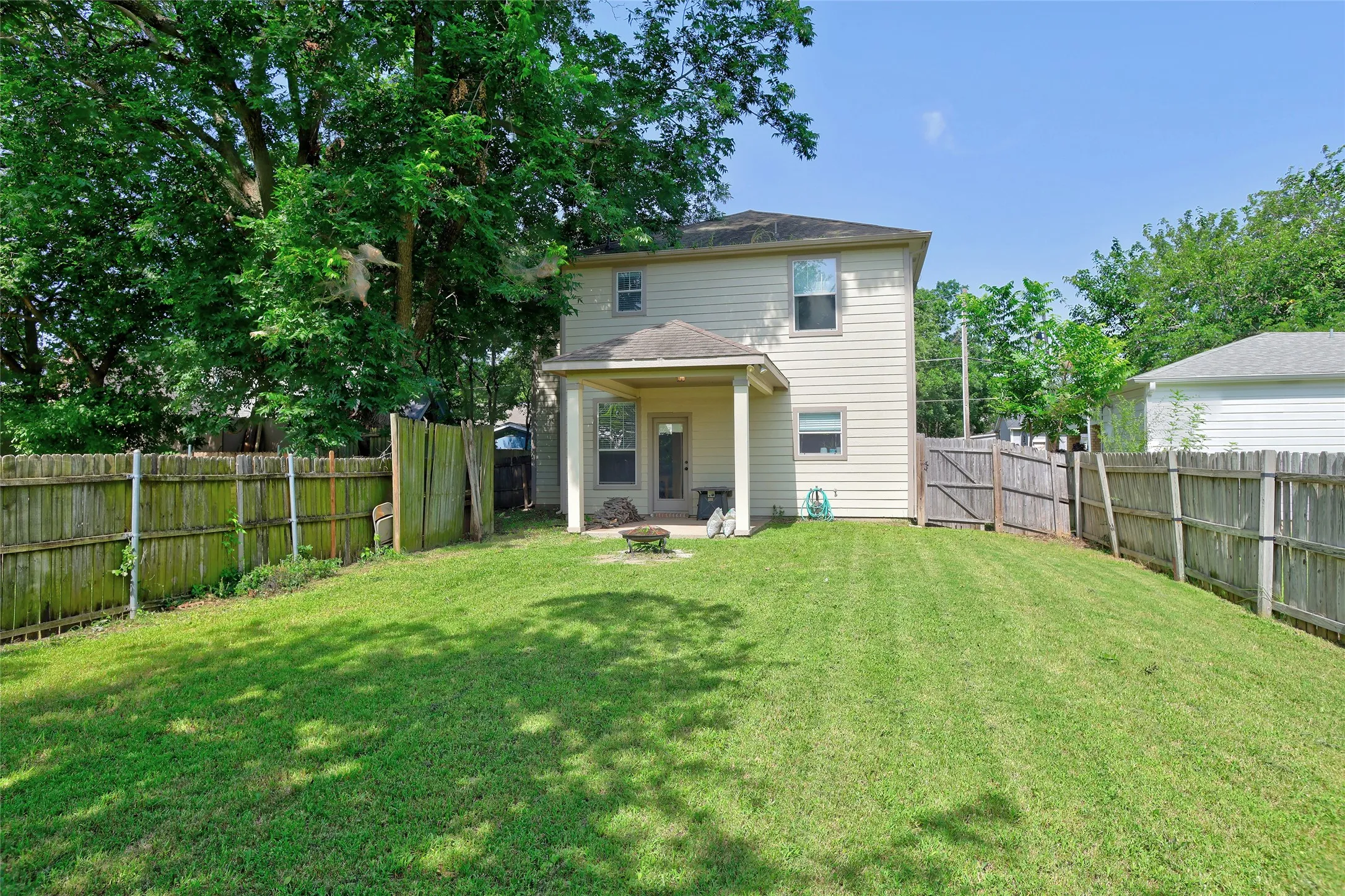 Expansive backyard featuring a large grass lawn, perimeter wood fencing, mature trees, and a covered patio area with a concrete slab