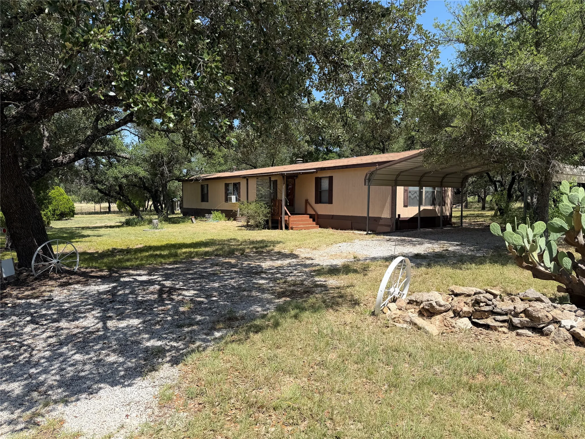 Single-story residence featuring a tan exterior with brown trim, covered front porch with wooden steps, an metal carport, a gravel driveway, and mature trees providing shade