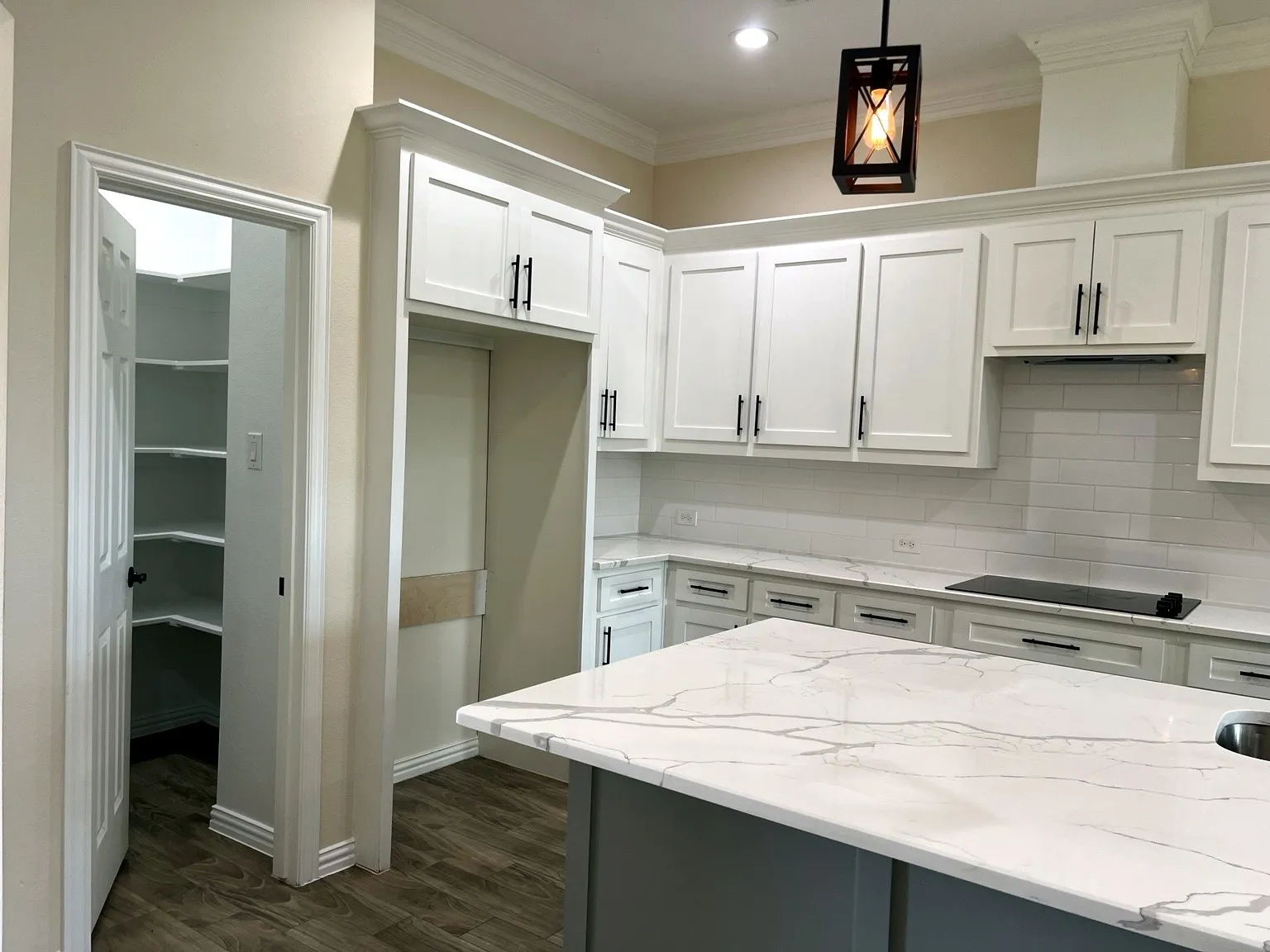 White shaker cabinetry with black hardware, white subway tile backsplash, white countertop island, wood-finish flooring, and a pantry with built-in shelving