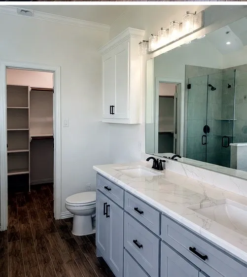 Dual vanity with quartz countertops, wood-finish flooring, a glass-enclosed shower, a built-in wall cabinet, and a walk-in closet with shelving
