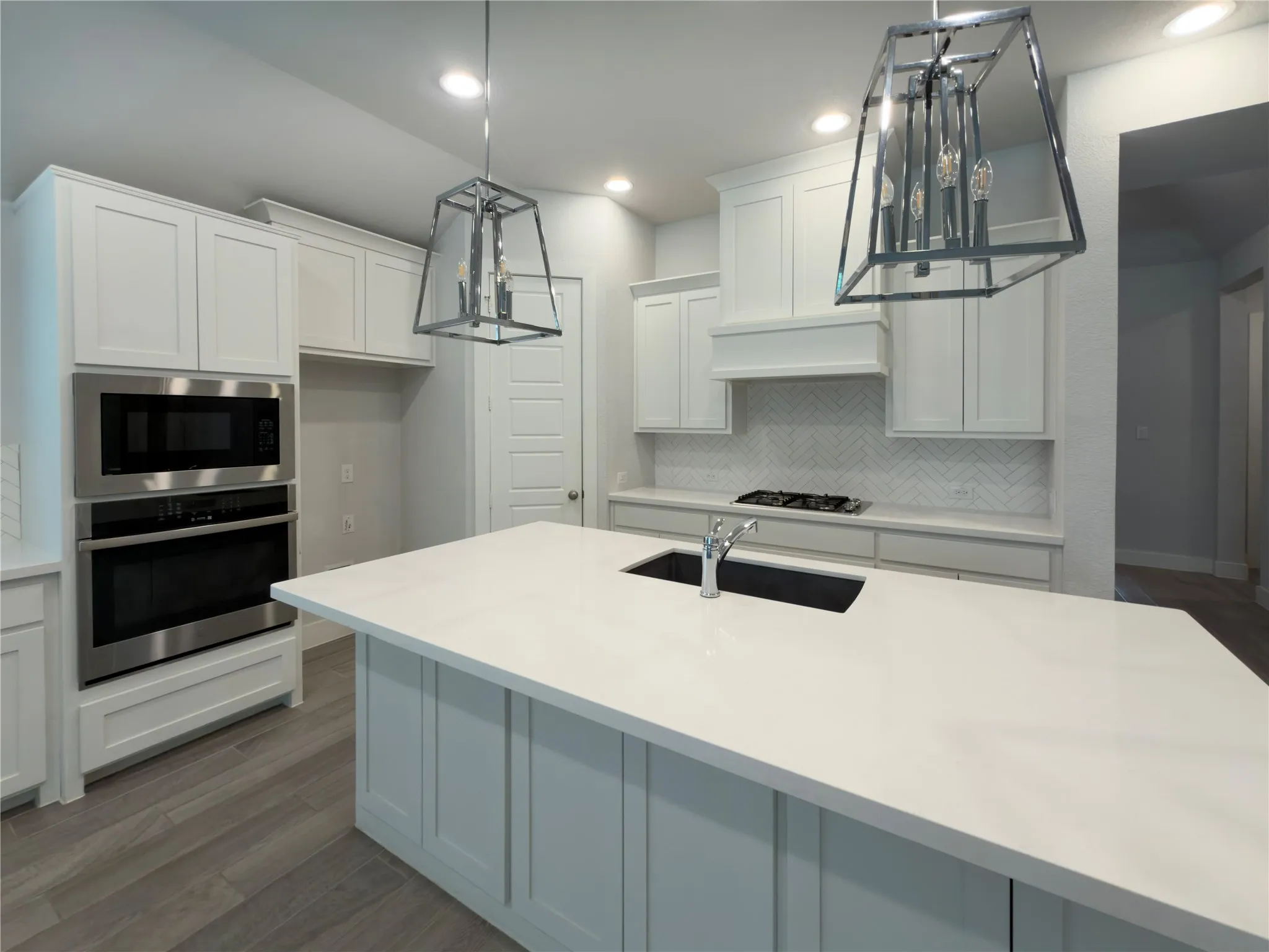 Kitchen featuring a large central island with an undermount sink, white shaker-style cabinetry, stainless steel built-in microwave and oven, a gas cooktop, and herringbone tile backsplash