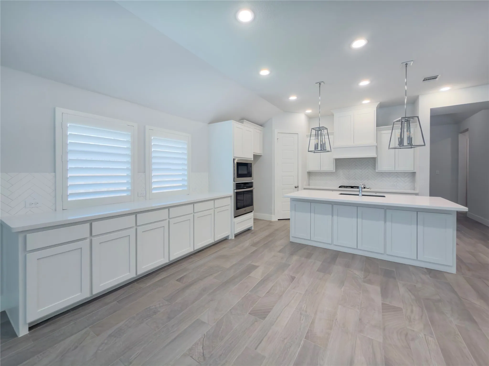 Modern kitchen featuring an expansive center island with integrated sink, white shaker-style cabinetry, stainless steel double wall ovens, wood-look plank flooring, and contemporary pendant lighting