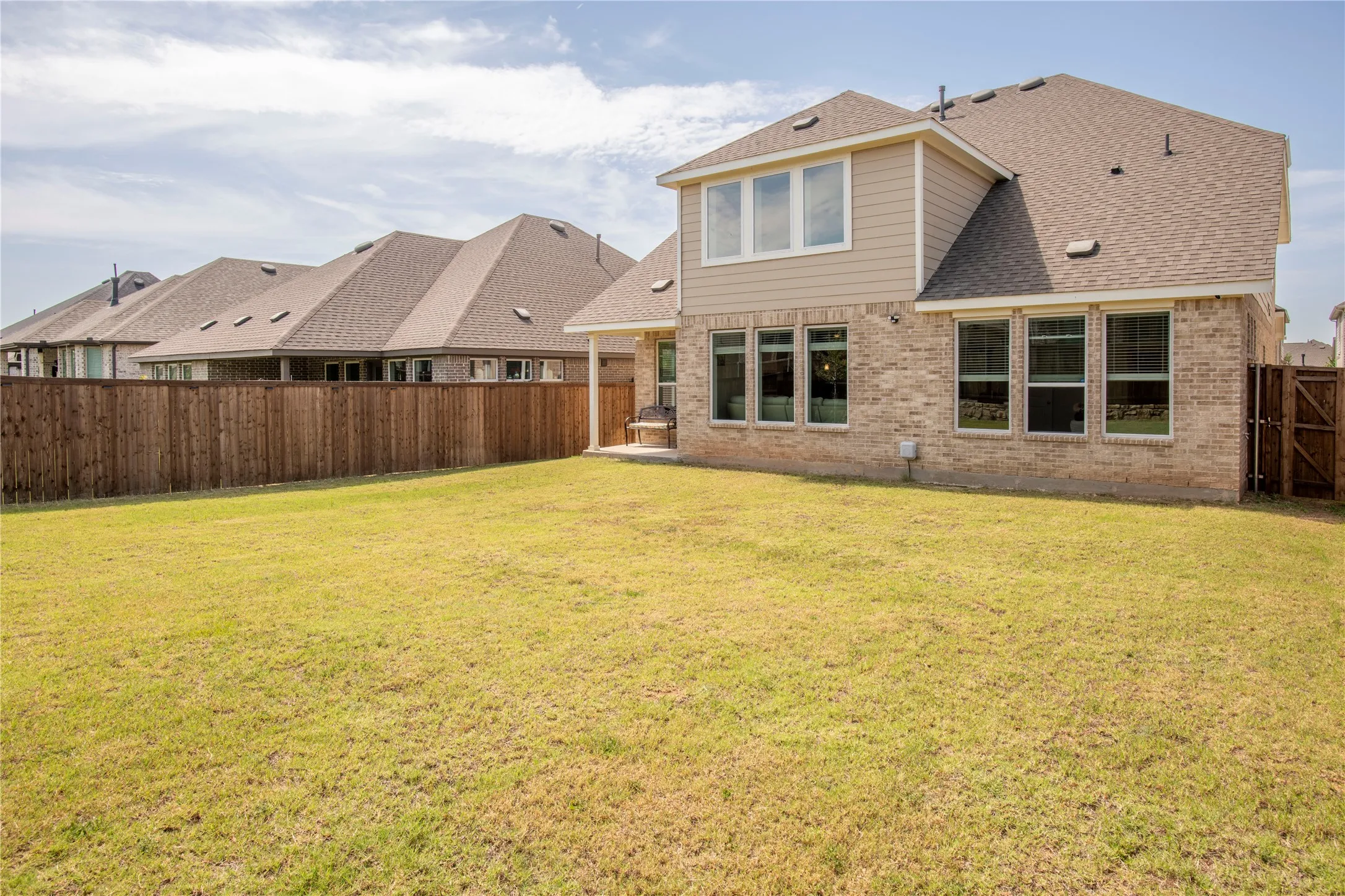 Expansive backyard featuring a privacy fence, a two-story residence with brick and siding exterior, multiple windows, and a covered patio area