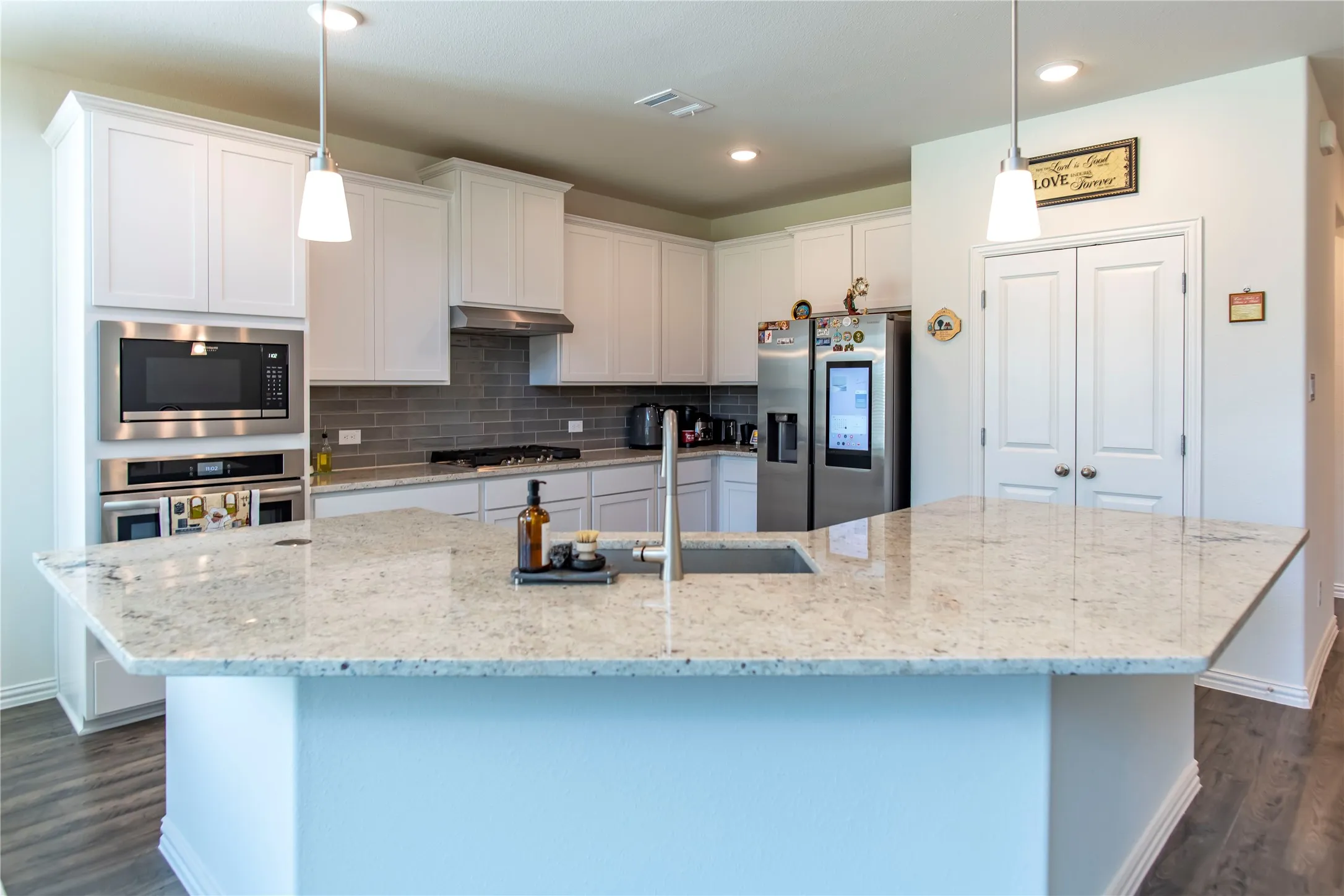 Kitchen featuring an expansive island with a light-toned countertop, white cabinetry, integrated stainless steel appliances, a gray subway tile backsplash, and wood-finish flooring