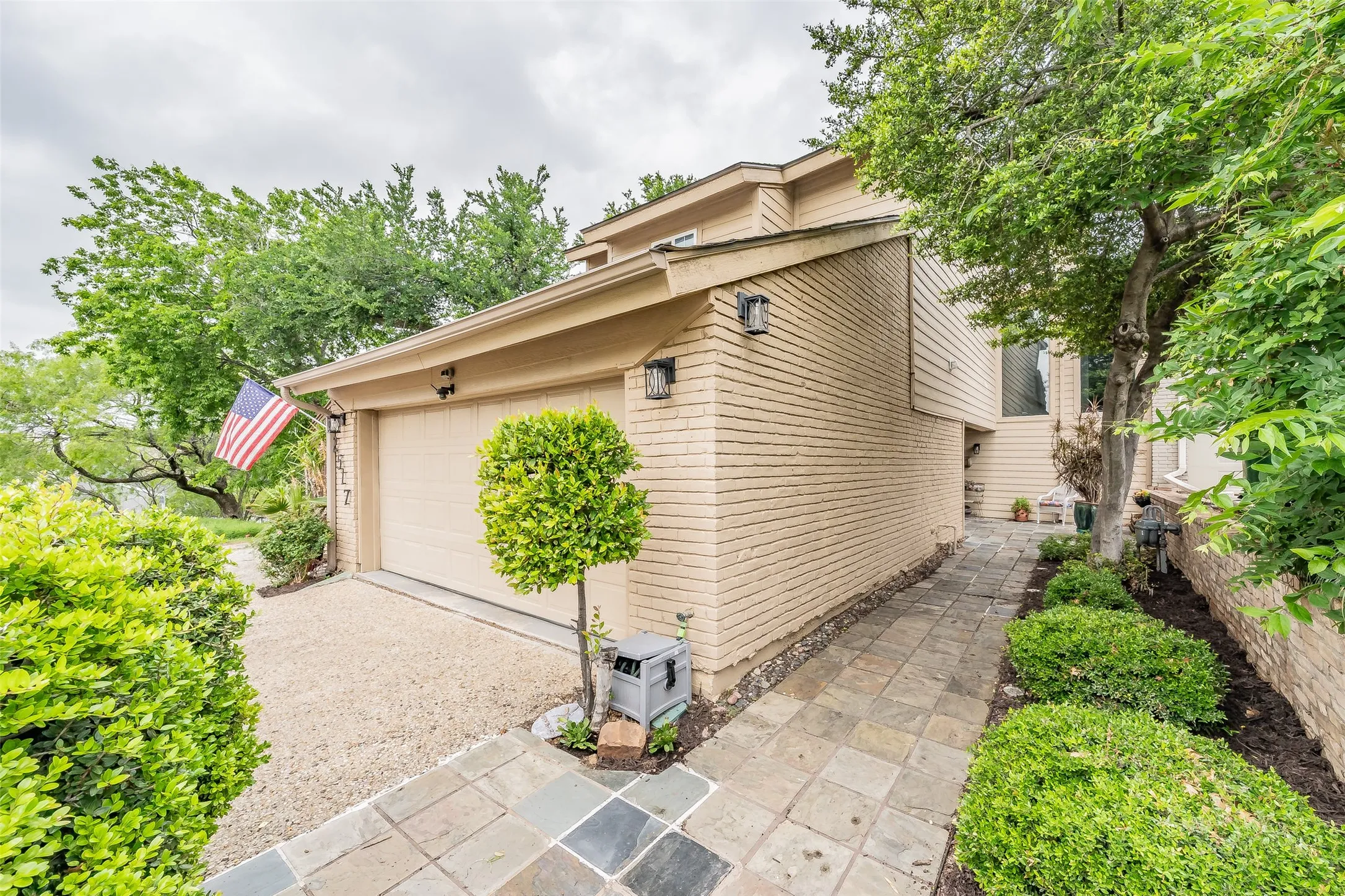 Property exterior featuring a single-car garage with a textured door, a slate tile walkway, mature landscaping, brick siding, and a gravel driveway