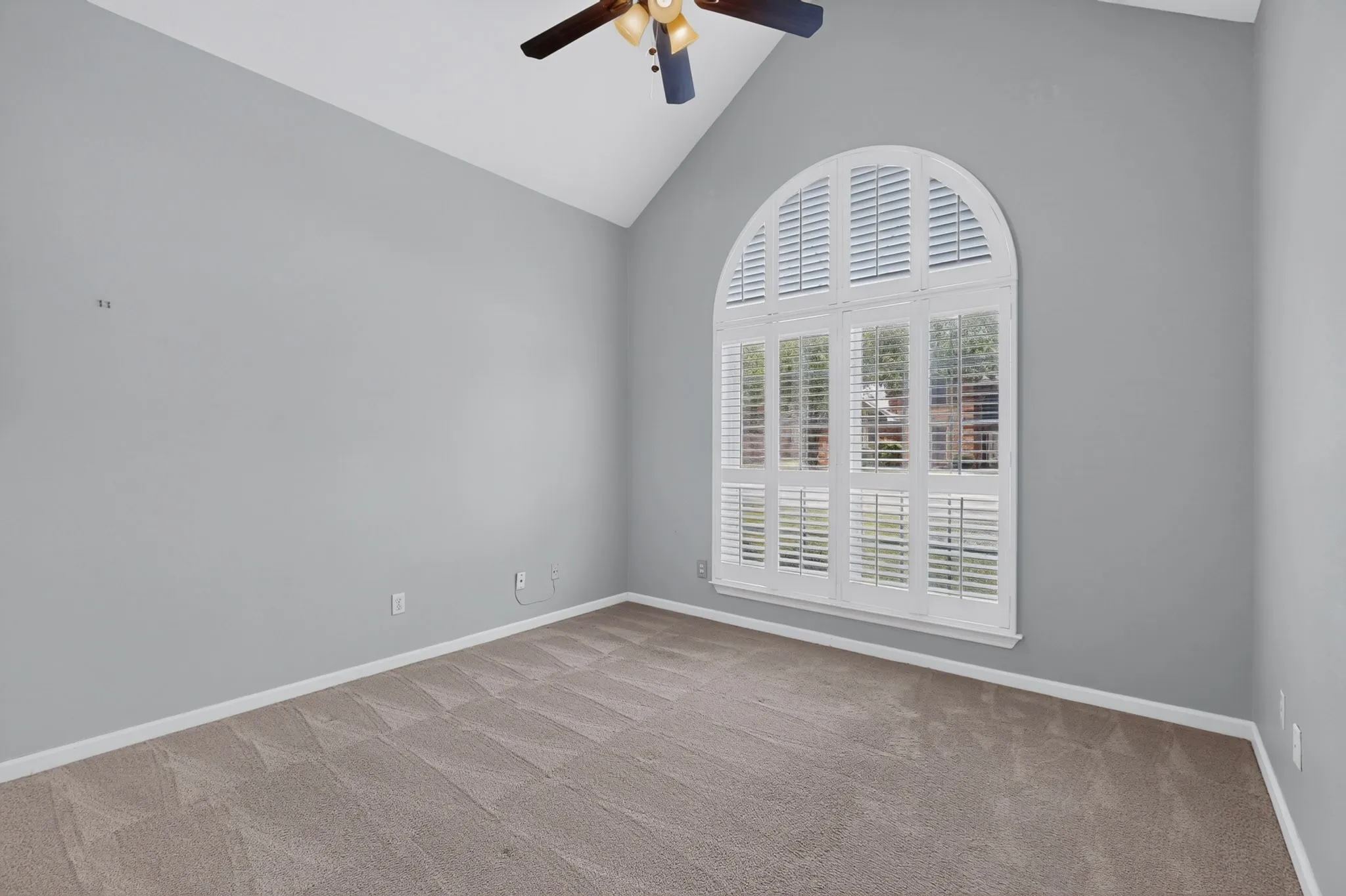 Arched window with white plantation shutters, vaulted ceiling, ceiling fan with light fixture, light gray wall color, and neutral carpet flooring