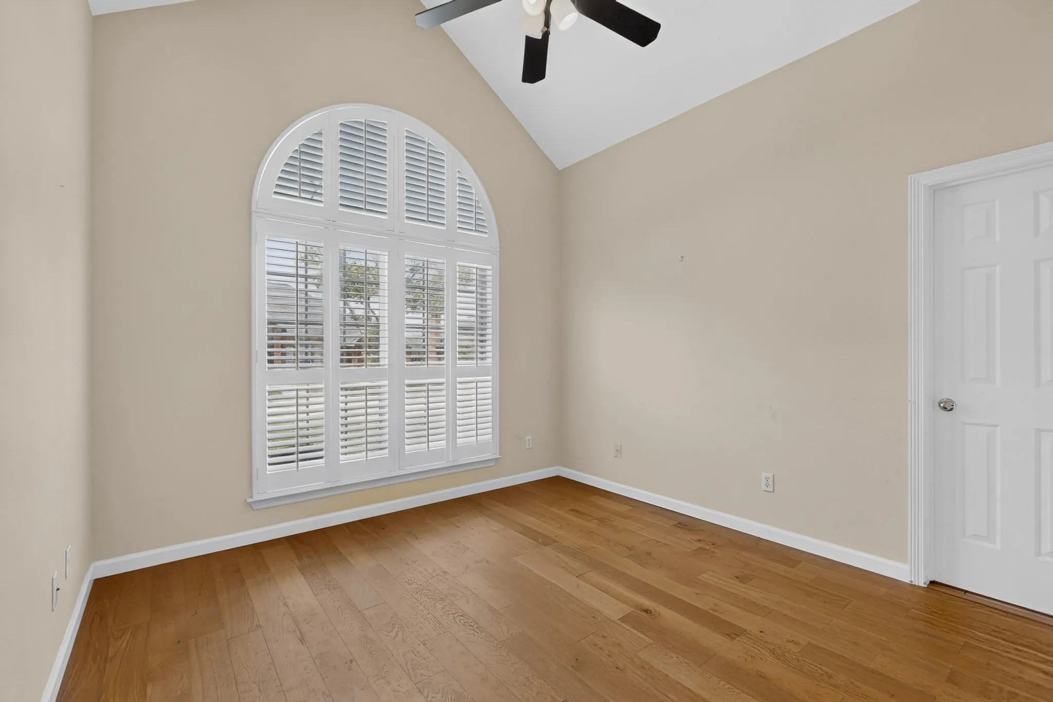 Vaulted ceiling room featuring a large arched window with white plantation shutters, wood-finish flooring, and a ceiling fan