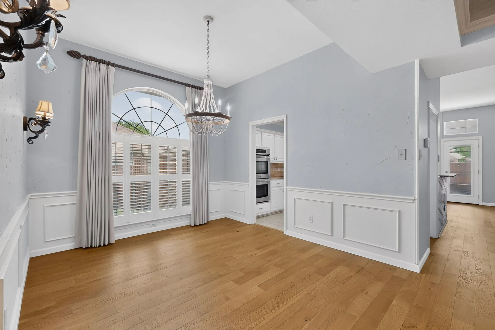 Spacious room featuring wood-finish flooring, wainscoting, and an arched window with plantation shutters