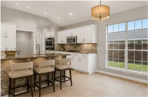 Kitchen featuring white cabinetry, stone-finish countertops, stainless steel appliances, and a stacked stone island with under-counter lighting. Virtually staged