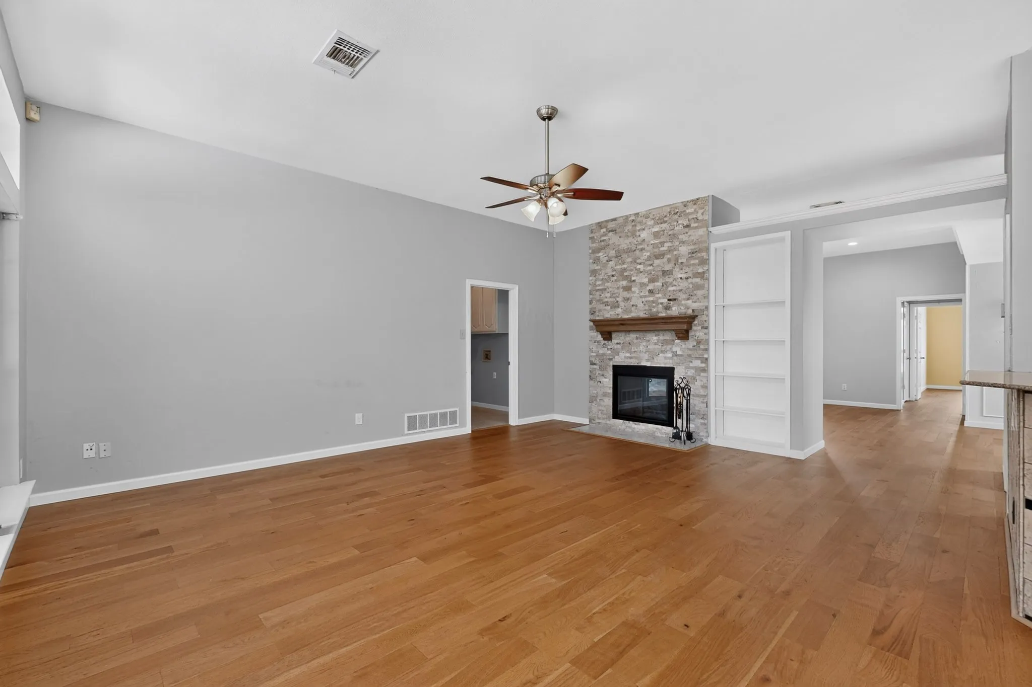 Spacious interior featuring wood-finish flooring, a stone-faced fireplace with a wood mantle, vaulted ceiling, a ceiling fan, and built-in shelving