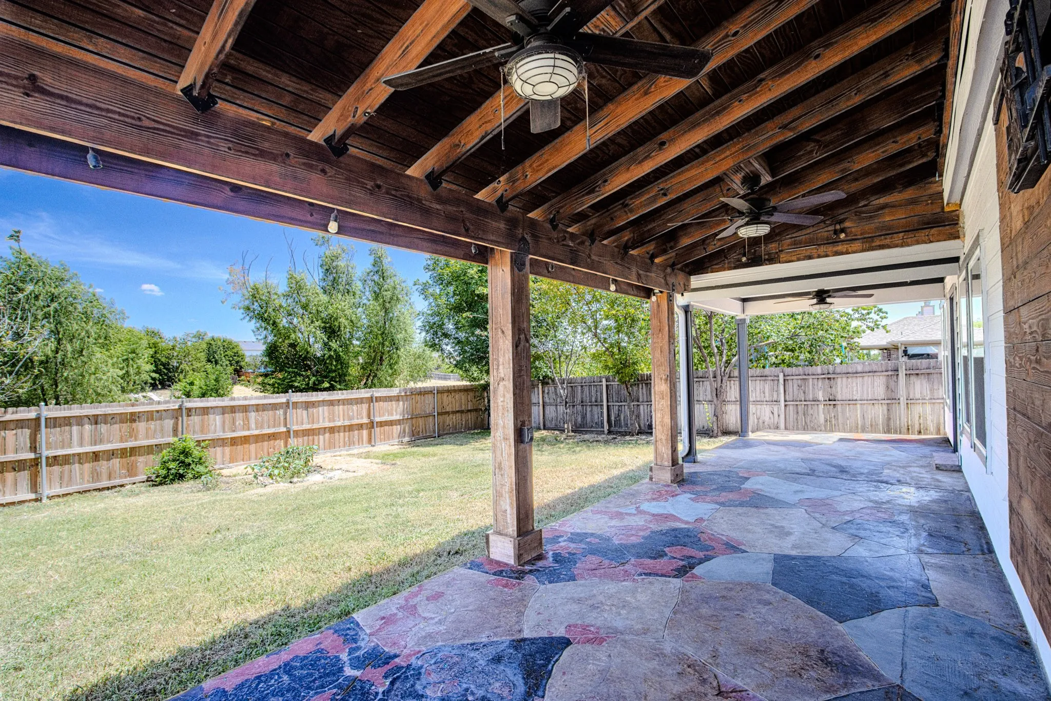 Expansive covered patio featuring a flagstone-style concrete surface, exposed wood beam ceiling, and two ceiling fans