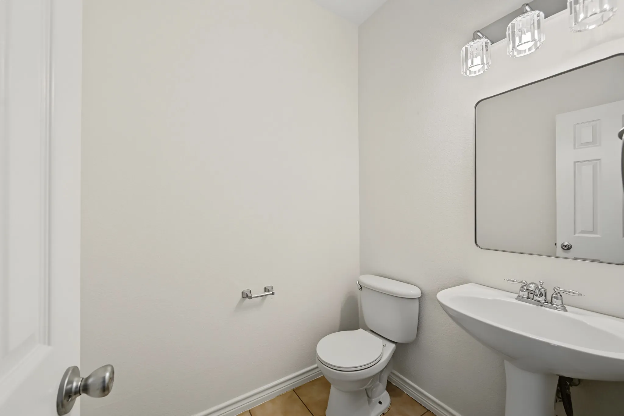 Pedestal sink with chrome faucet, a toilet, a wall-mounted mirror, and a three-light vanity fixture