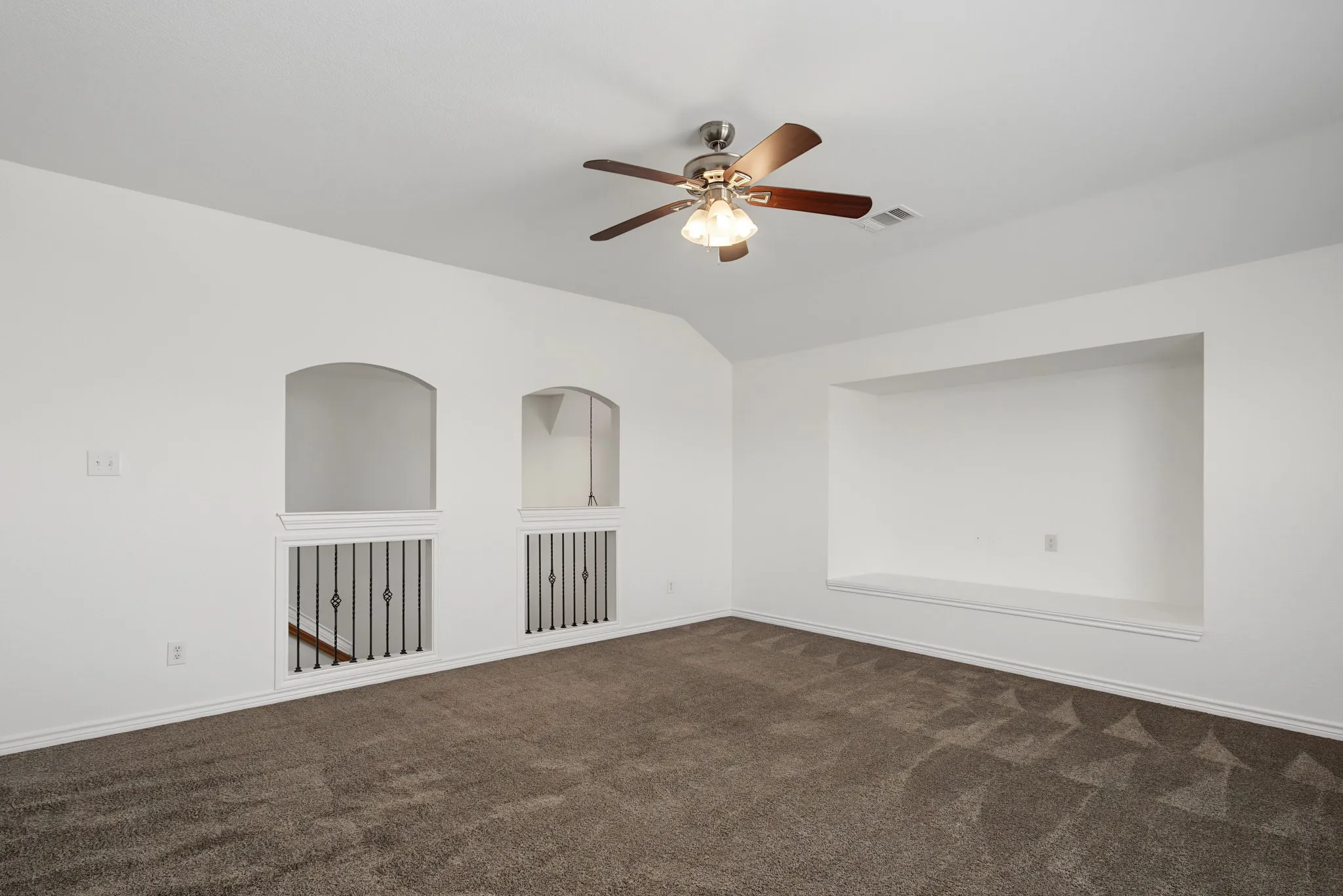 Carpeted room featuring a ceiling fan with light kit, a built-in wall niche, and arched cutouts with iron balusters overlooking a stairway