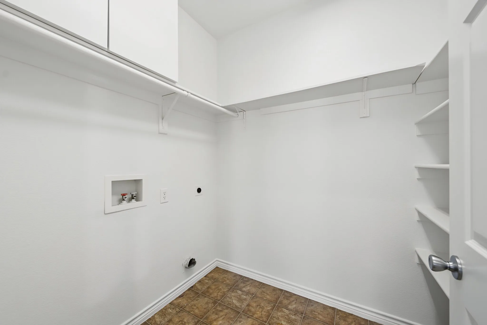 Laundry room featuring white shelving, a wall-mounted hanging rod, built-in water hookups, and patterned brown tile flooring