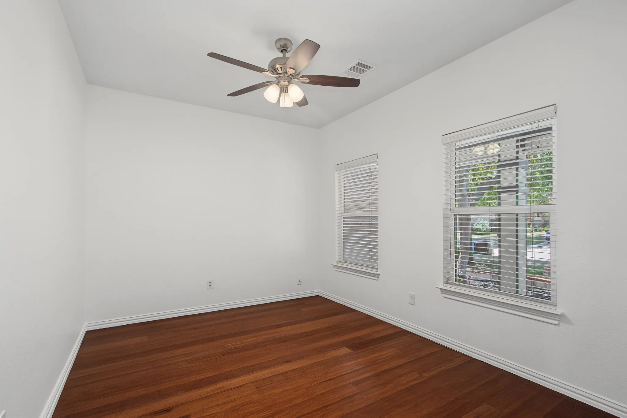 Spacious room featuring wood-finish flooring, two windows with blinds, a ceiling fan with integrated lighting, and white baseboards