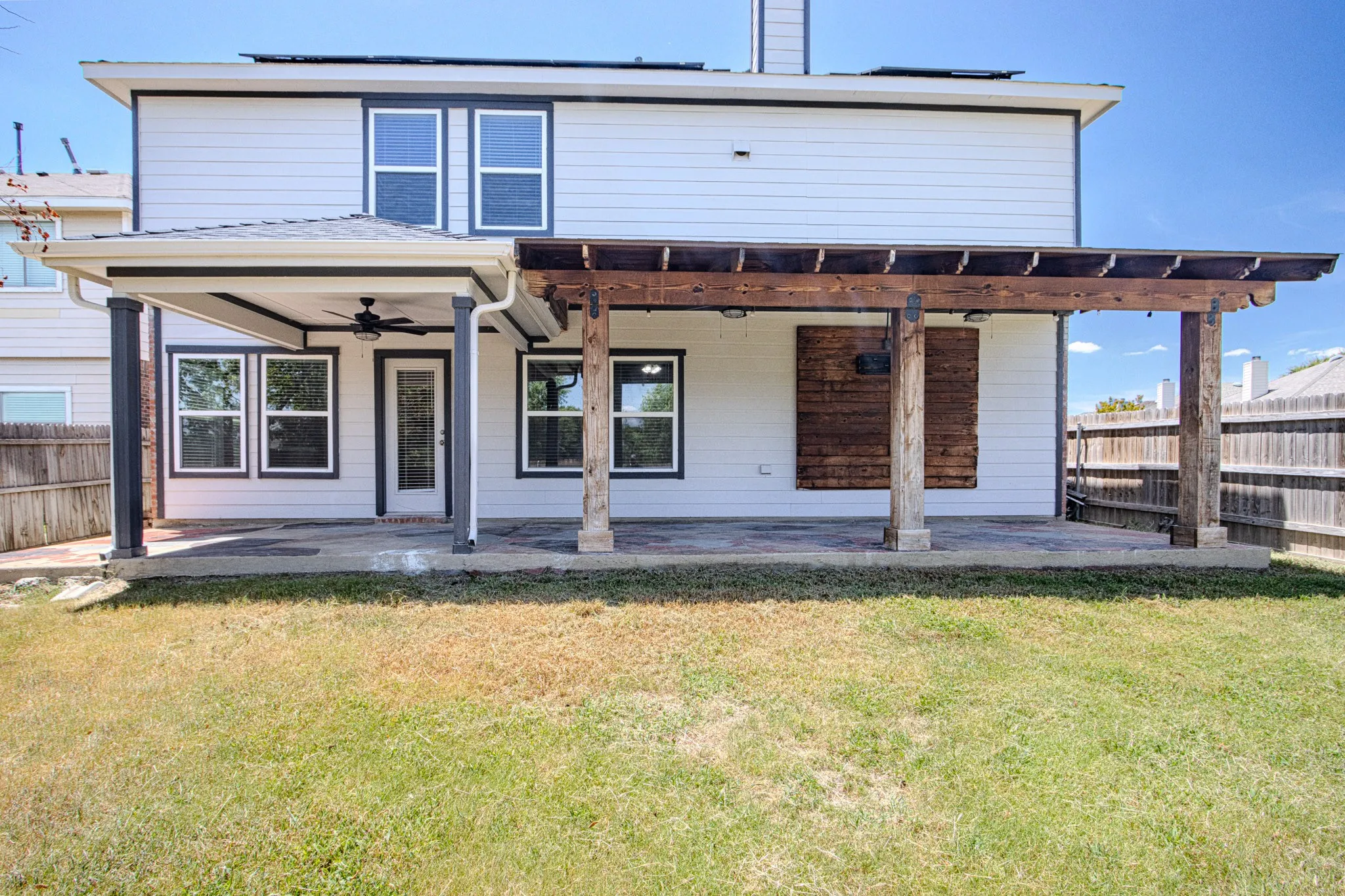 Rear exterior featuring a covered patio with a ceiling fan, an adjacent pergola, a glass-paneled door, multiple windows, and a wood privacy fence
