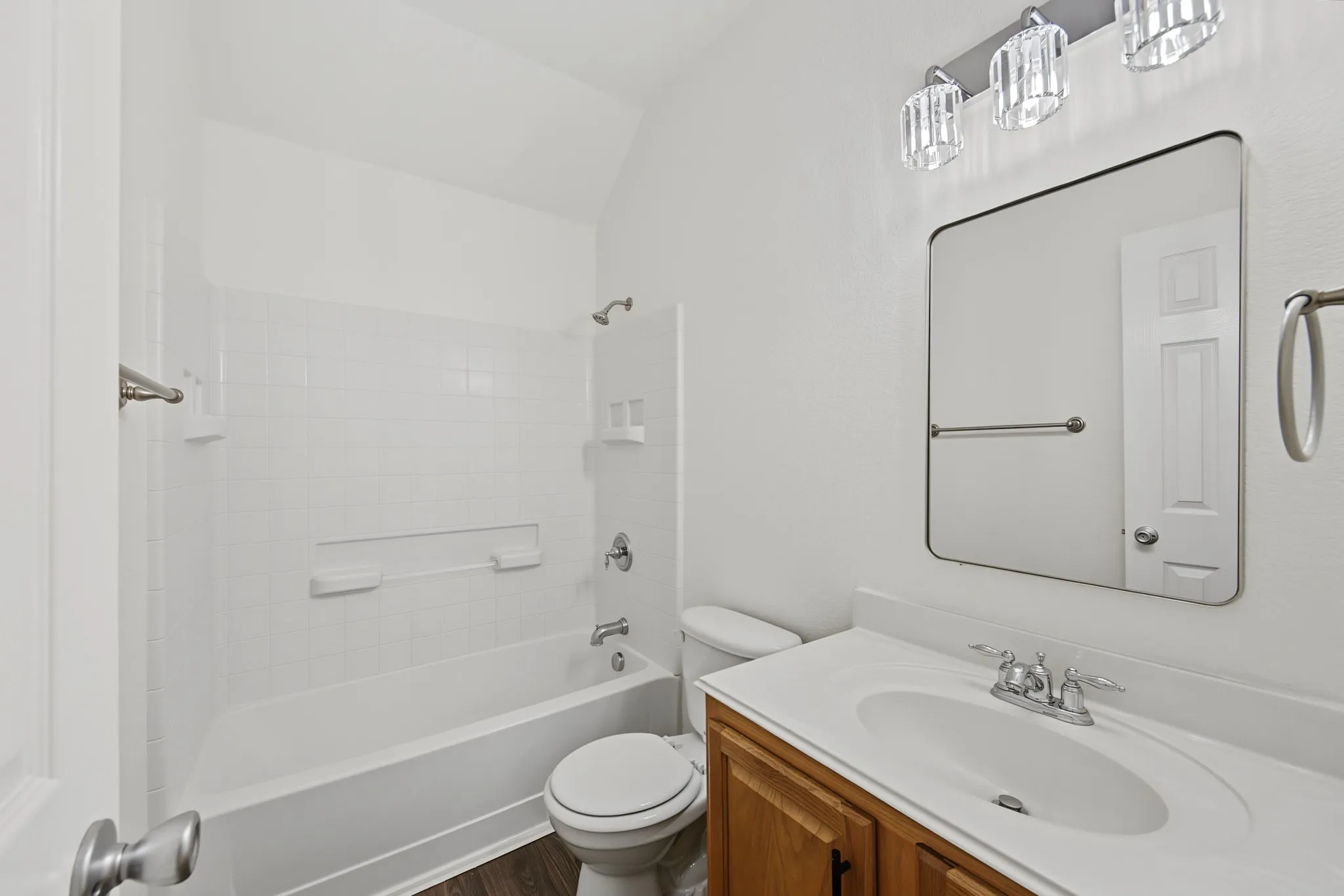 Bathroom featuring a white bathtub, tiled shower surround, wood-finish vanity with an integrated sink, and a rectangular mirror with a three-light vanity fixture