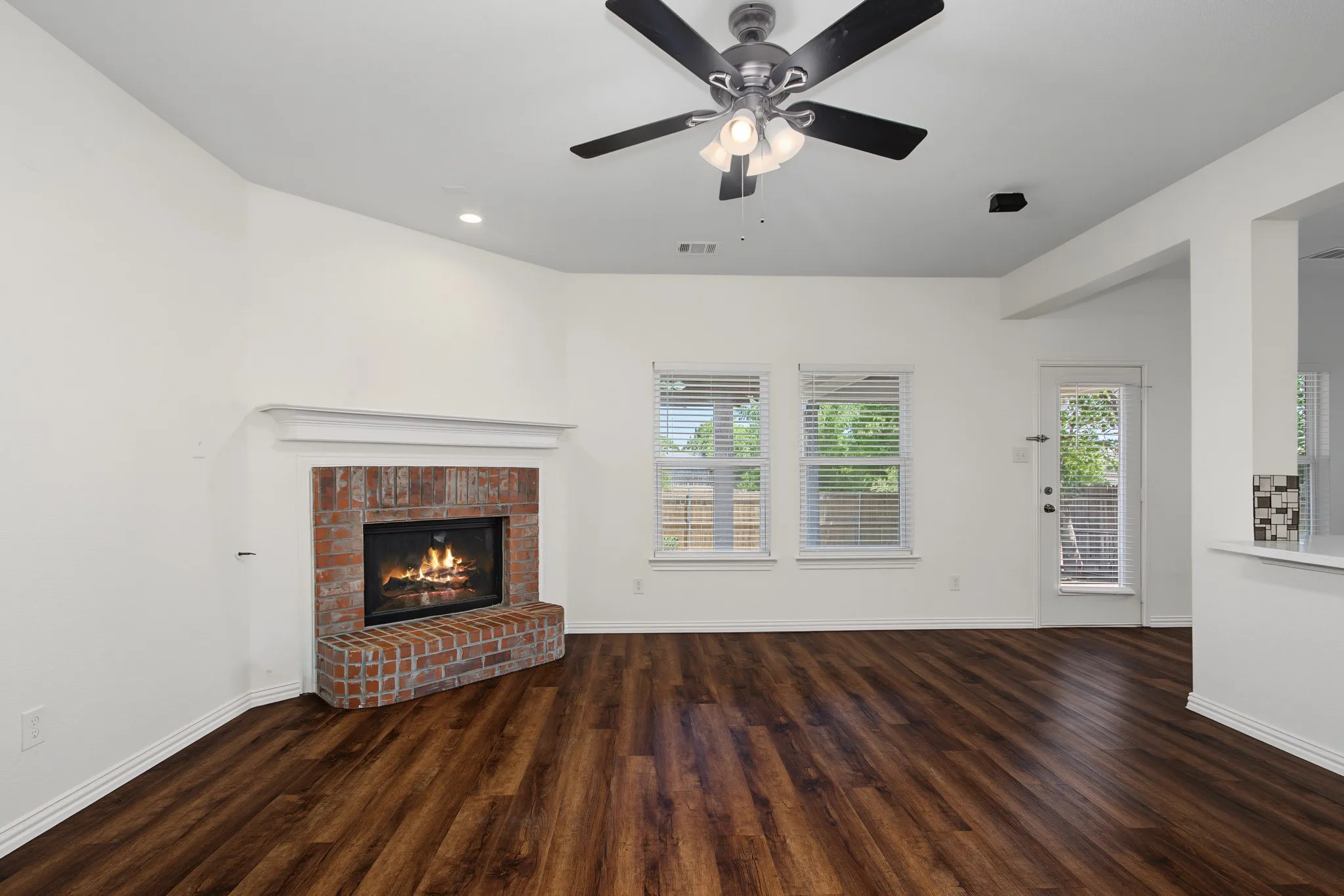Spacious living area featuring a corner brick fireplace with a white mantel, wood-finish flooring, and multiple windows with blinds