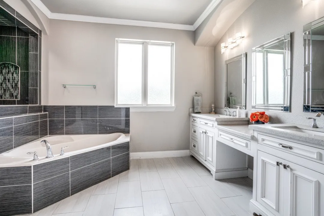 Bathroom featuring a corner soaking tub with dark tile surround, dual white vanity cabinets with integrated sinks, light-toned countertop, and light tile flooring