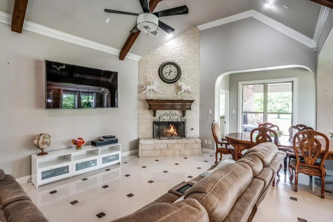 Vaulted ceiling with exposed wood beams and a ceiling fan, a stone fireplace with a wood mantel, light-toned flooring with dark square inlays, and an arched entryway to an adjacent room with a window overlooking exterior greenery
