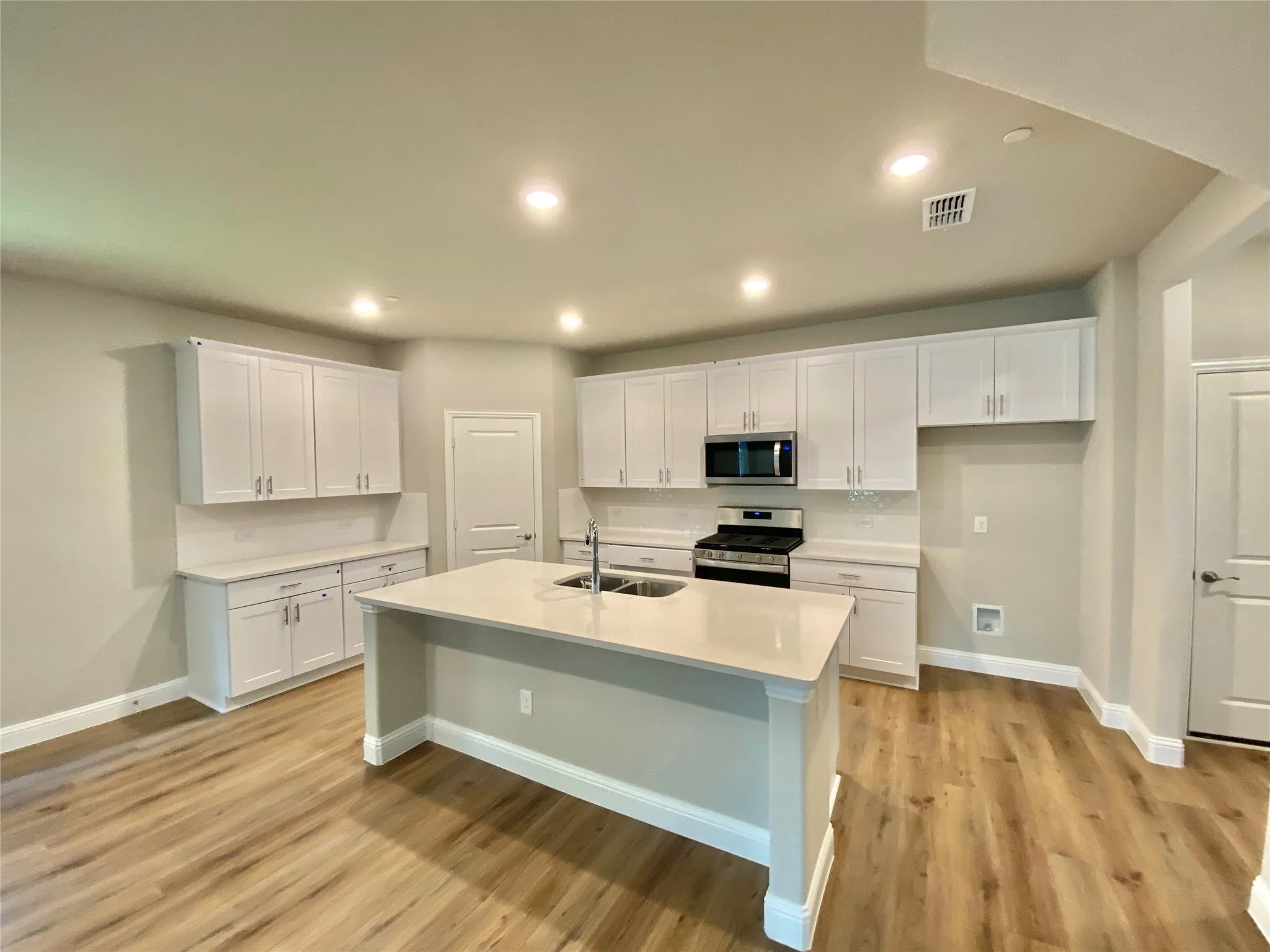 Kitchen featuring an expansive island with a double basin sink, white cabinetry with brushed nickel hardware, light-toned countertops, and wood-finish flooring