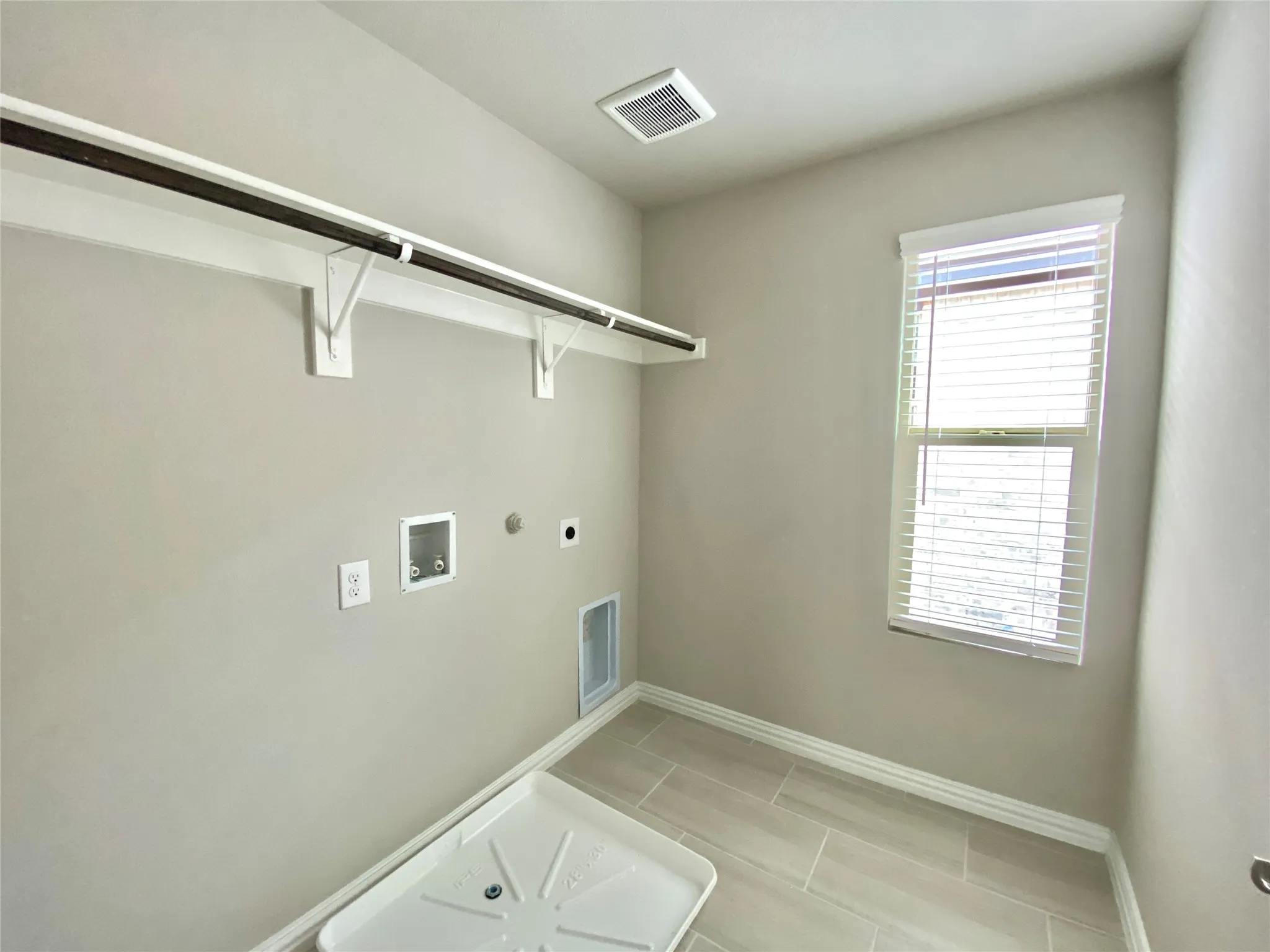 Dedicated utility space featuring a window with horizontal blinds, overhead ventilation, a clothing rod with shelving, wall-mounted electrical outlets, and light-toned tile flooring