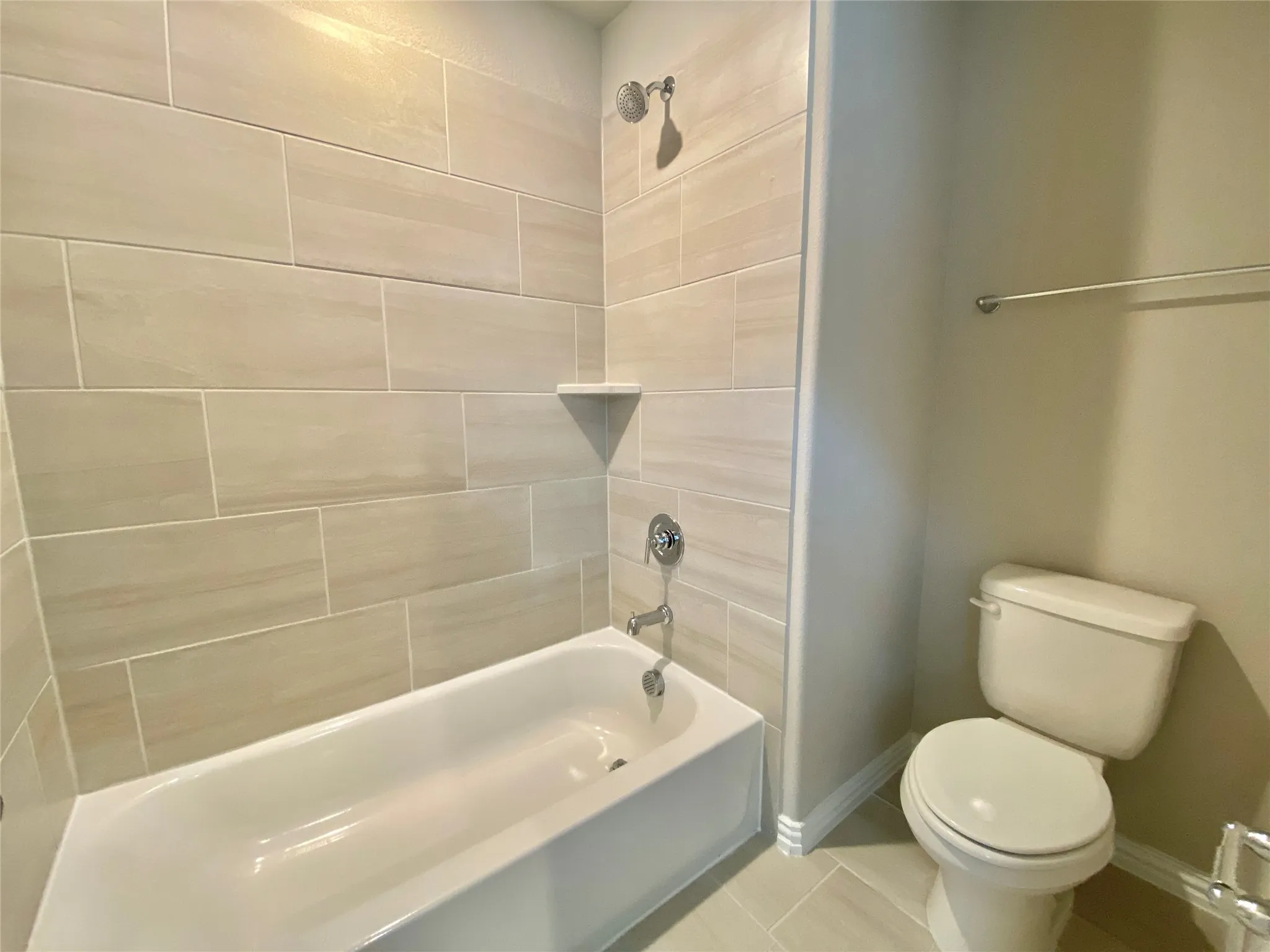 Bathroom featuring a white tub with chrome fixtures, light-toned rectangular tile surround, and a corner shelf