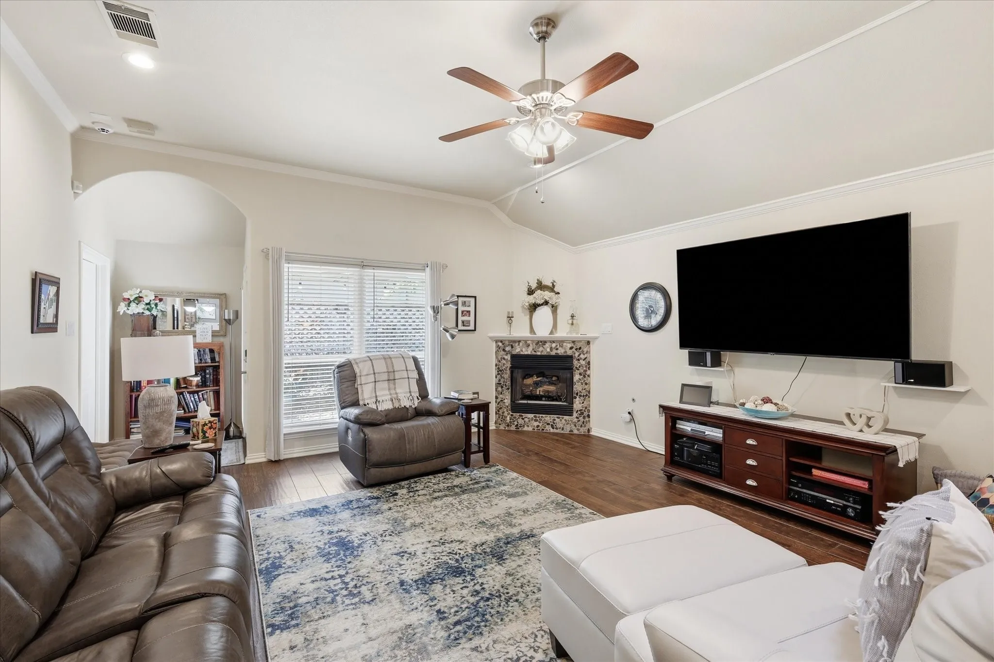 Living room featuring wood-finish flooring, a ceiling fan with integrated lighting, and a corner fireplace with a tiled surround