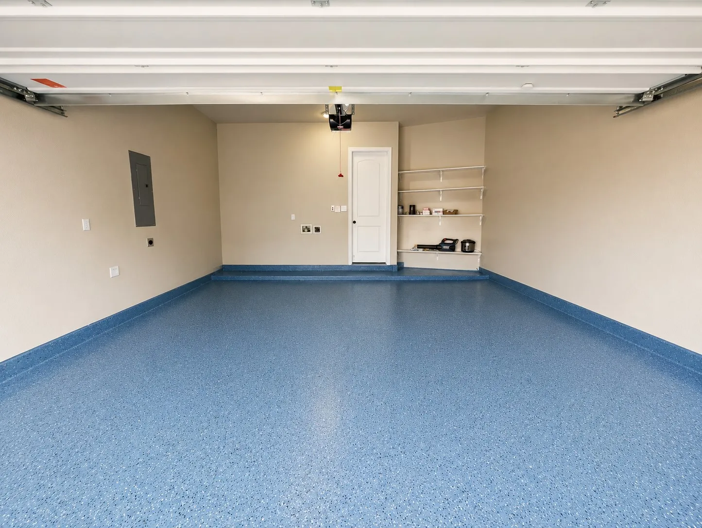 Spacious garage featuring a speckled blue epoxy floor coating, light beige walls, built-in shelving, an interior access door, and a visible electrical panel