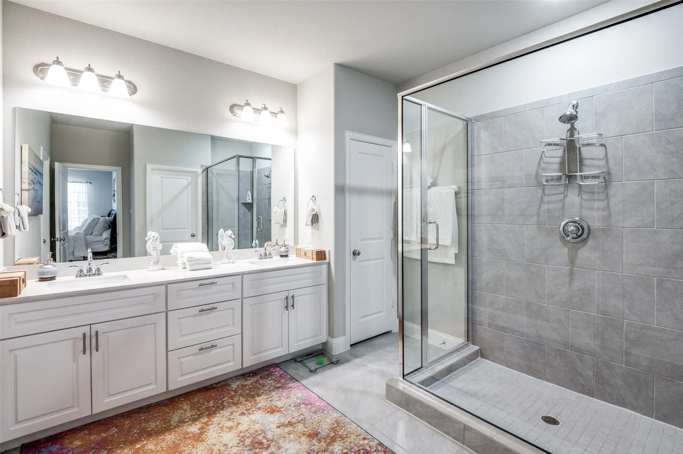 Bathroom featuring a dual vanity with white cabinetry and chrome fixtures, extensive mirror with overhead lighting, glass-enclosed shower with gray subway tile surround, and a separate door