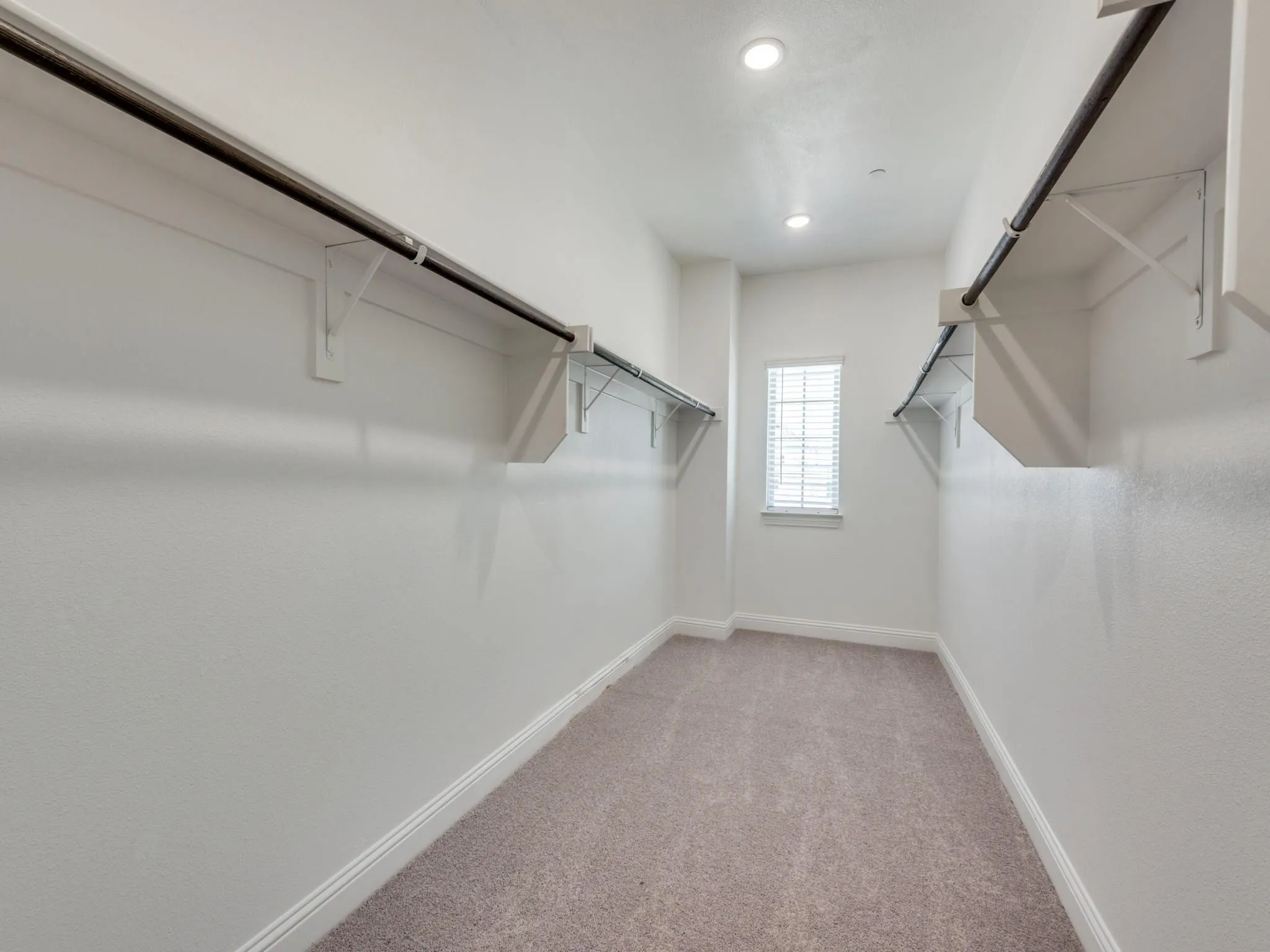 Spacious closet with ample hanging rods, integrated shelving, recessed lighting, a window with blinds, and neutral carpet flooring