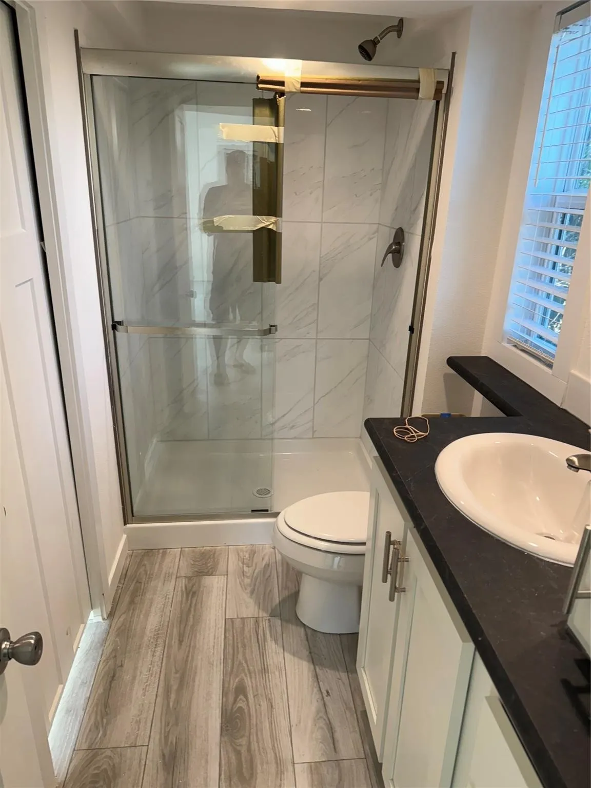 Full bathroom featuring a glass-enclosed shower with tile surround, white vanity with integrated sink and dark countertop, wood-finish plank flooring, and a window with blinds