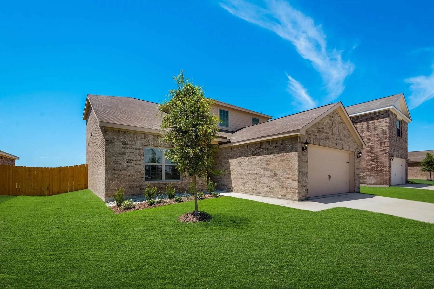 Brick exterior residence featuring an attached garage with a tan door, a paved driveway, and a front yard with a young tree and landscaping