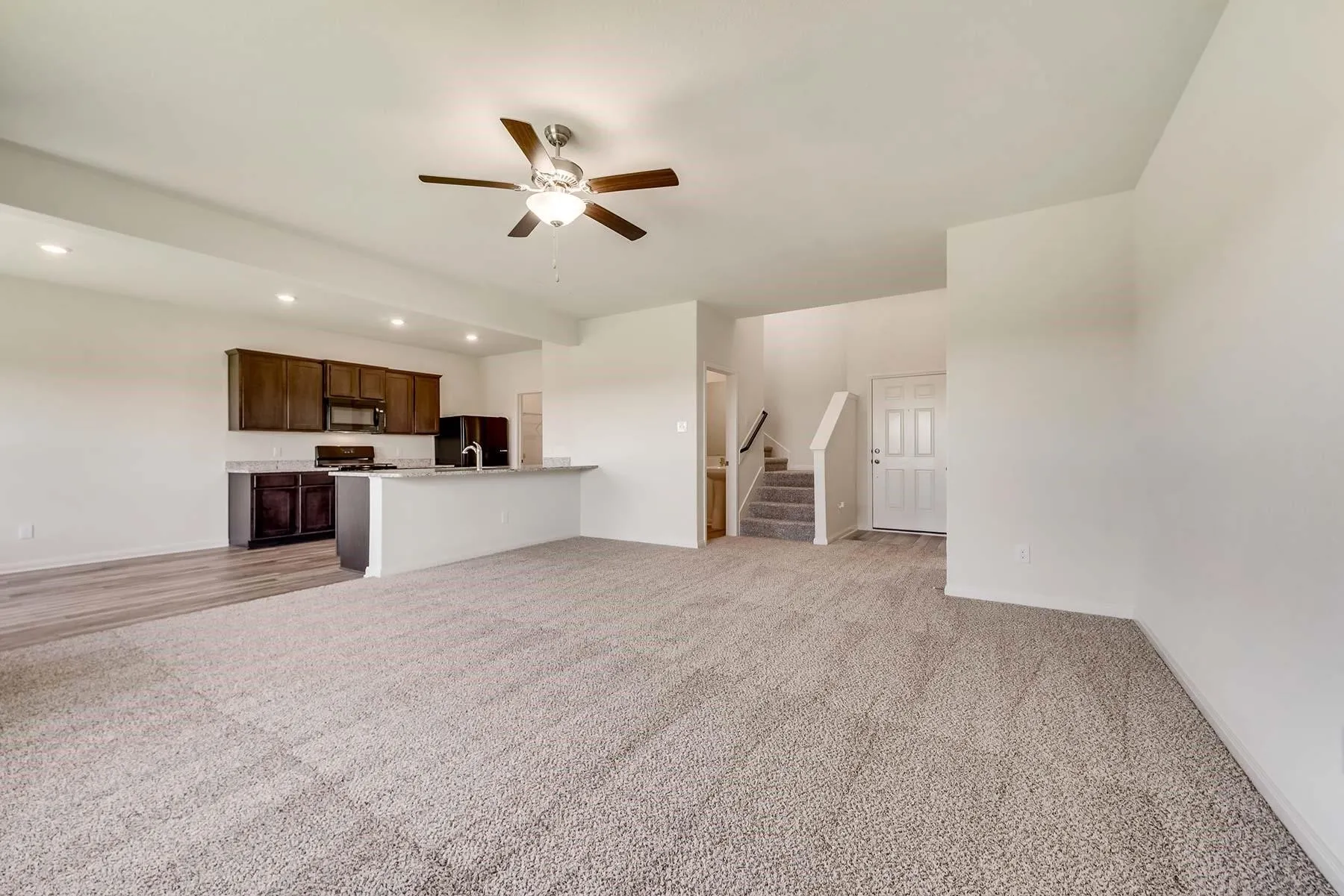 Open-concept living area featuring light-toned carpeting, a kitchen with dark wood cabinetry, a breakfast bar with a light-colored countertop, and a ceiling fan with integrated lighting