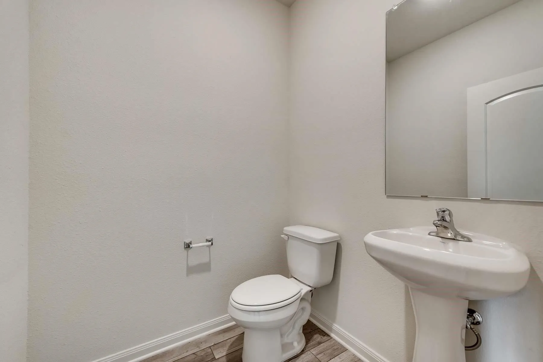Bathroom featuring a pedestal sink, chrome faucet, wall-mounted mirror, white toilet, and wood-finish flooring with white baseboards