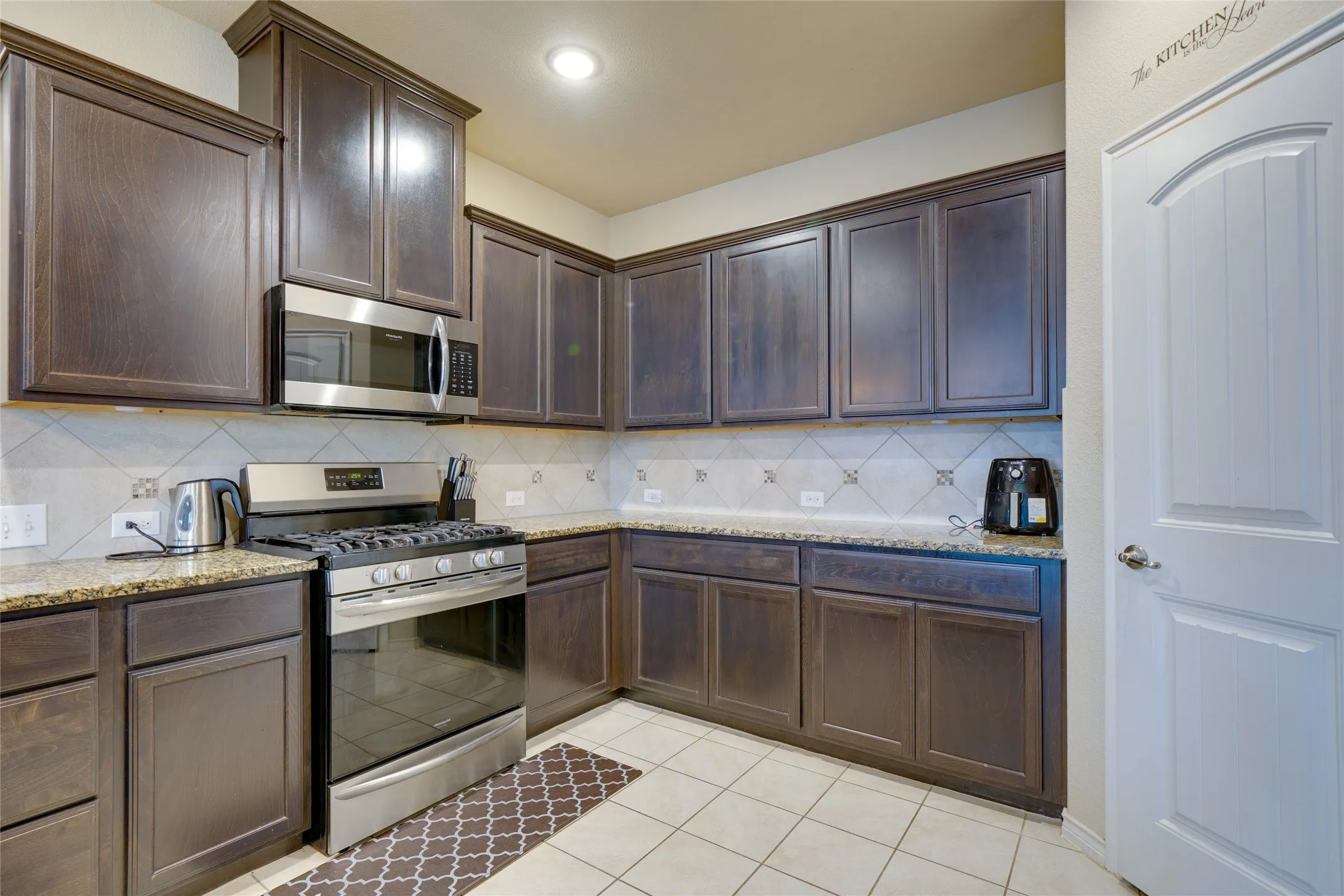 Kitchen with appliances with stainless steel finishes, tasteful backsplash, light stone counters, light tile patterned floors, and dark brown cabinetry