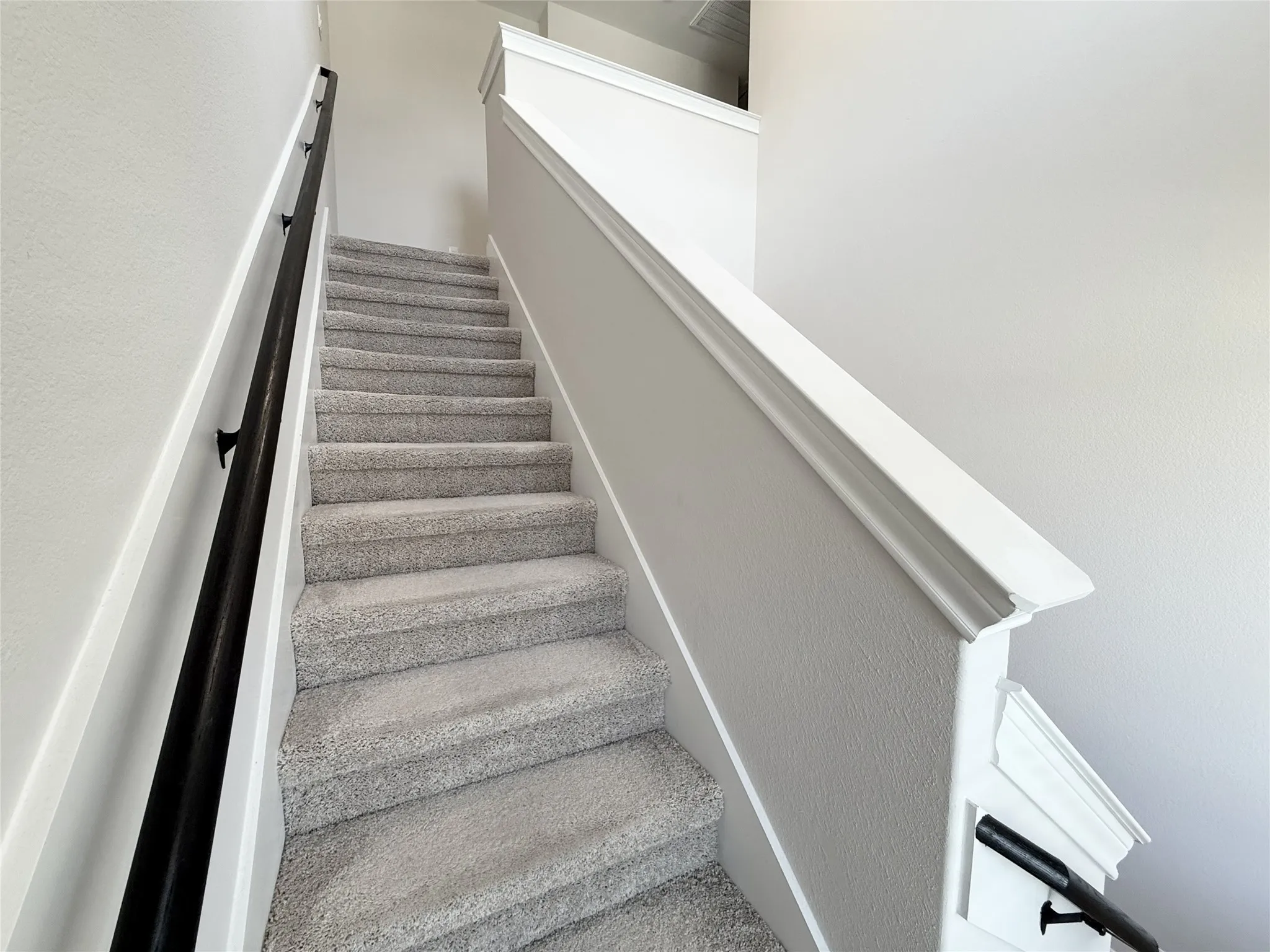 Staircase featuring light gray carpeting, crisp white risers, a contrasting black wall-mounted handrail, and white decorative trim